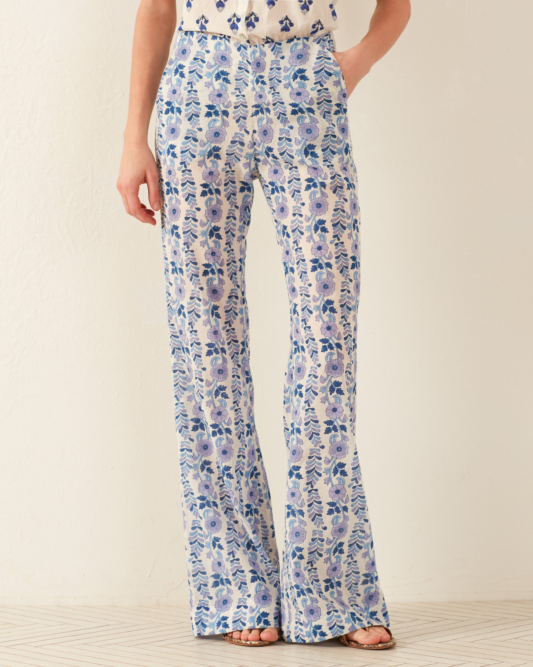 A person is wearing the Charlie Iris Ivy Pant, featuring a wide-leg floral print in shades of blue and white. It is paired with a white top that showcases a hand-block printed design. The backdrop consists of a neutral off-white wall and light-colored floor, offering an elegant contrast to the vibrant attire.