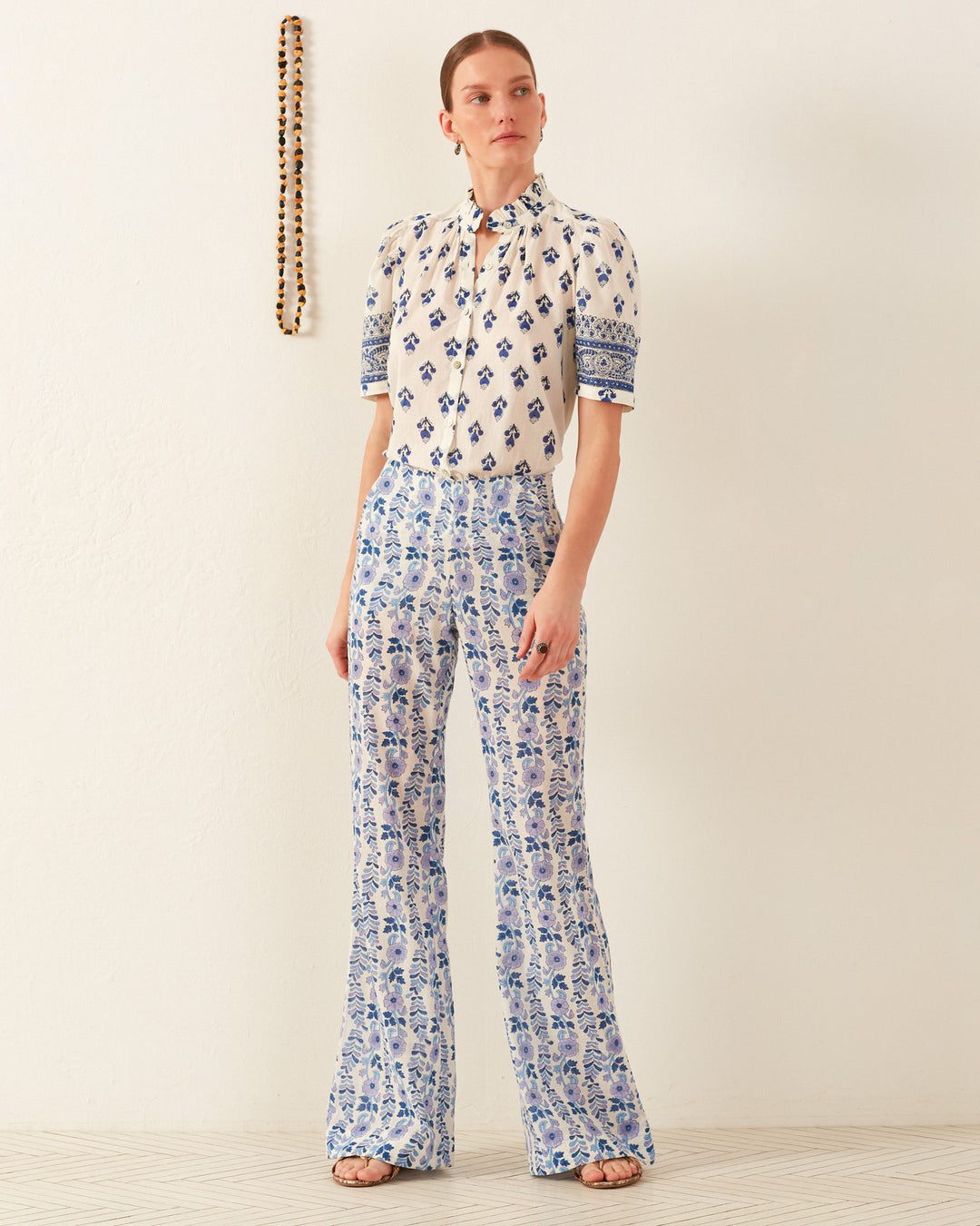 A person stands against a light wall, wearing a patterned blouse and Charlie Iris Ivy Pant featuring hand-block printed floral designs in shades of blue. A decorative string with a trailing ivy motif hangs on the wall. The person is barefoot and looking slightly to the side.