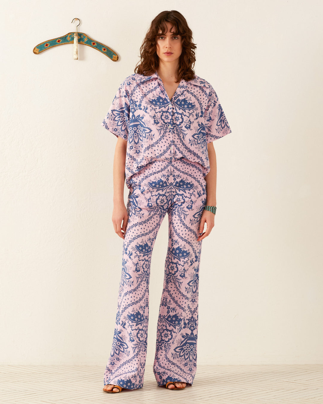 A person stands against a plain white background, wearing a matching pink and blue patterned shirt with the Charlie Oasis Bandana Pant, known for its retro flared design. A decorative hanger is mounted on the wall behind them. The person has curly hair and is wearing open-toed shoes.