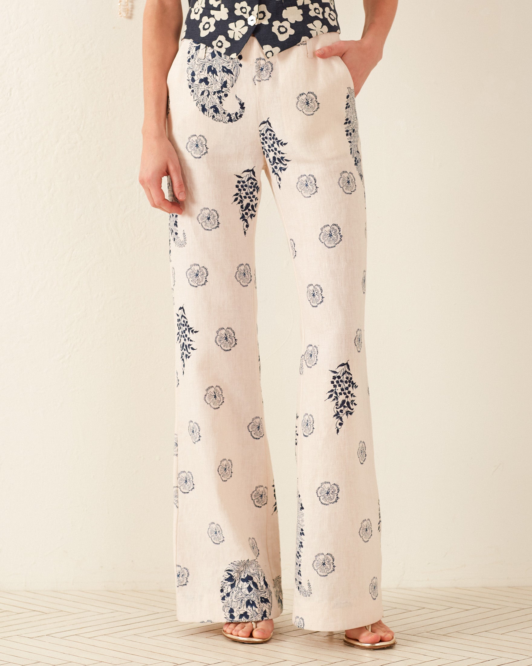 A person wearing the Charlie Porcelain Linen Pant, which showcases a '70s-inspired floral motif in a cream color with wide legs. Their hand is tucked into a pocket, and they are paired with open-toed sandals, standing on a floor with a subtle geometric design.