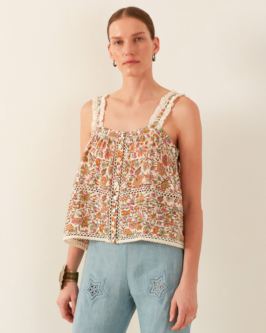 A person stands against a plain background wearing the Effie Garden Party Silk Top, paired with light blue pants adorned with star patterns. Their hair is tied back and accessorized with bracelets and earrings, adding an elegant touch to the ensemble.