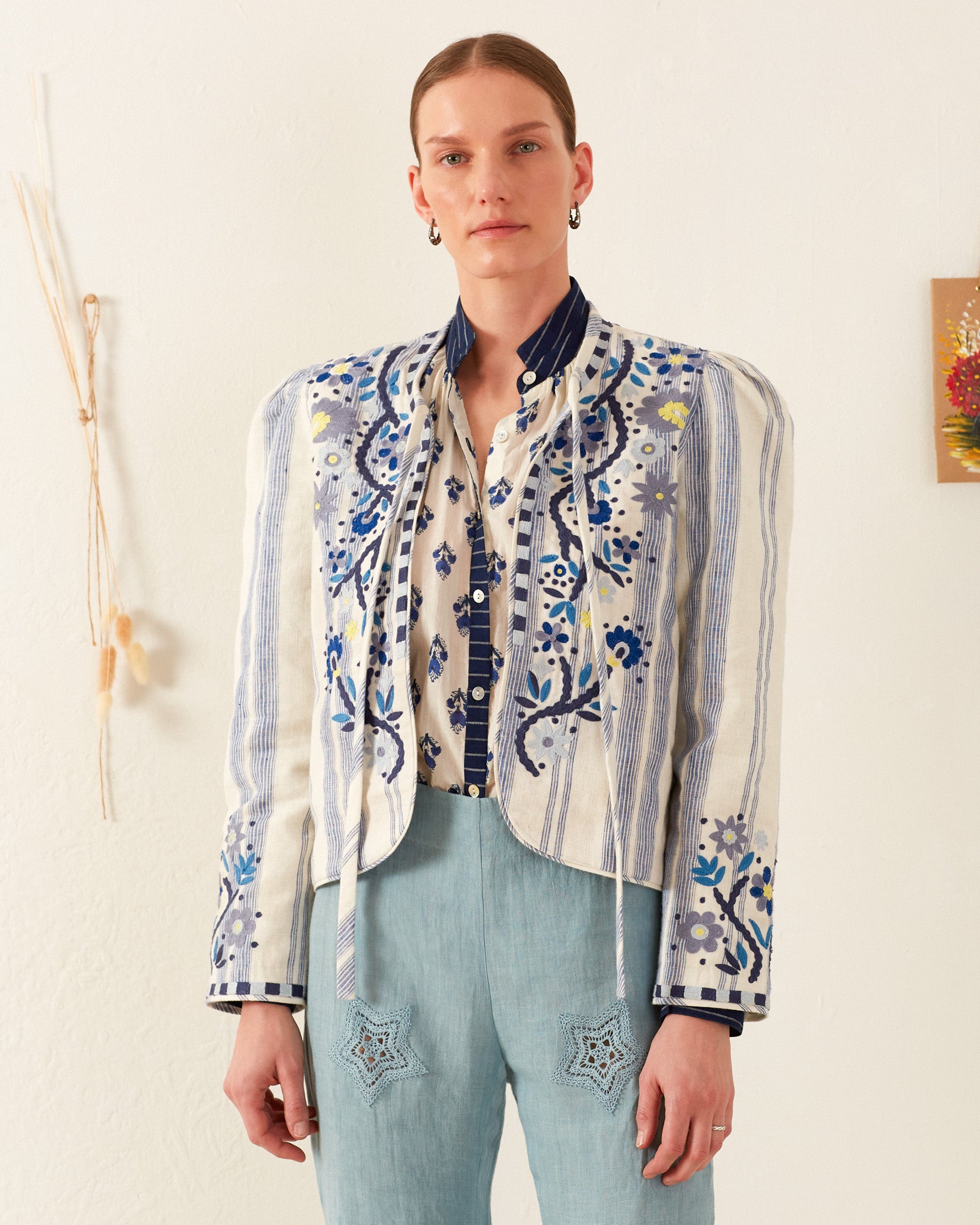 A person is posing against a plain wall, showcasing the Fiorello Pacific Reef Jacket, which features intricate blue floral embroidery and vertical stripes. They wear a patterned shirt underneath, and light blue pants decorated with star motifs. To finish off the look, they have styled their hair casually and are wearing earrings.