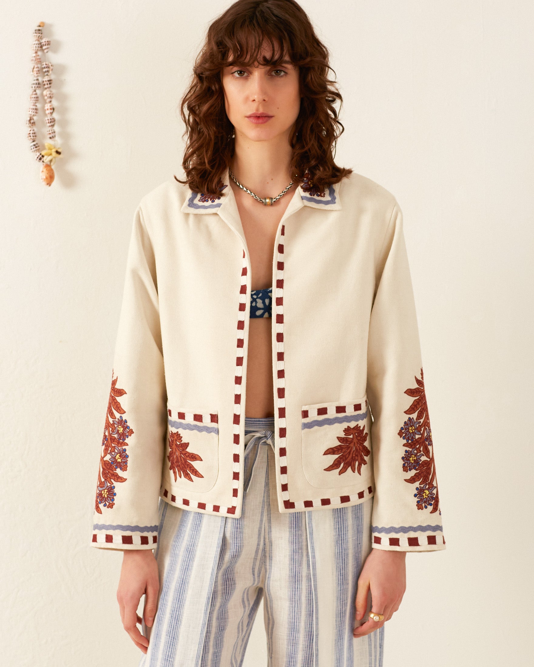 A person stands against a plain wall wearing the Francis Waterlily Bloom Jacket, characterized by its folk-inspired design and floral patterns on the sleeves and pockets, over a blue patterned top. They also wear hand-embroidered striped pants. A beaded decoration hangs on the wall to the left.