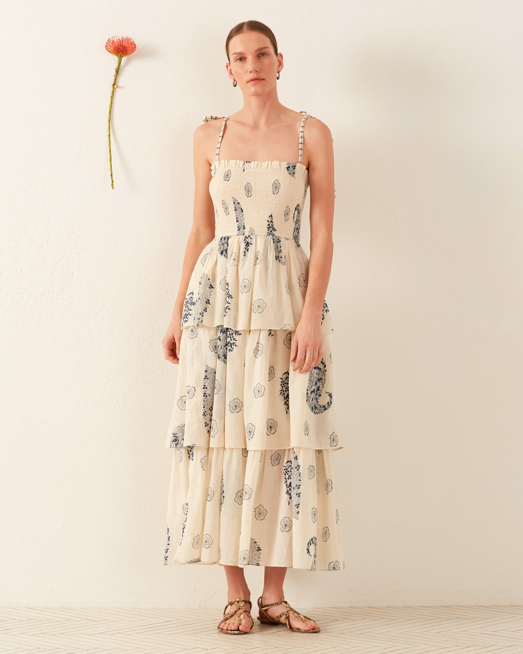 A model poses against a light background wearing the Kaia Porcelain Tier Dress, featuring a sleeveless design and blue floral print. Made from hand-loomed cotton, she stands barefoot with a single flower affixed to the wall beside her.