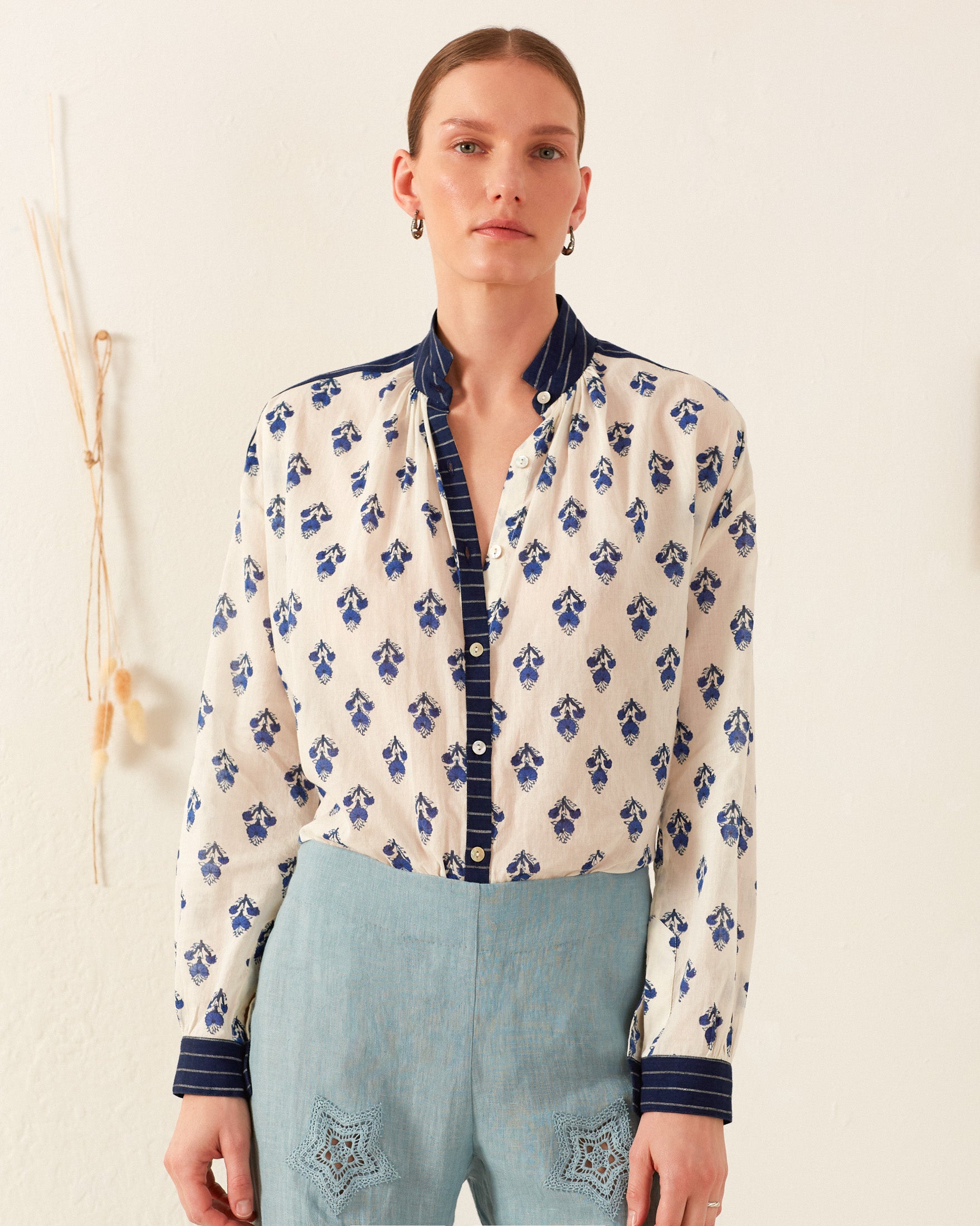 A person stands against a cream-colored wall, wearing the Kiki Indigo Bhutti Shirt paired with turquoise pants. The shirt features a white base with a blue floral pattern and dark blue collar and cuffs. Their hair is tied back, and they accessorize with hoop earrings, exuding the charm of block-printed designs. Simple decor is visible in the background.