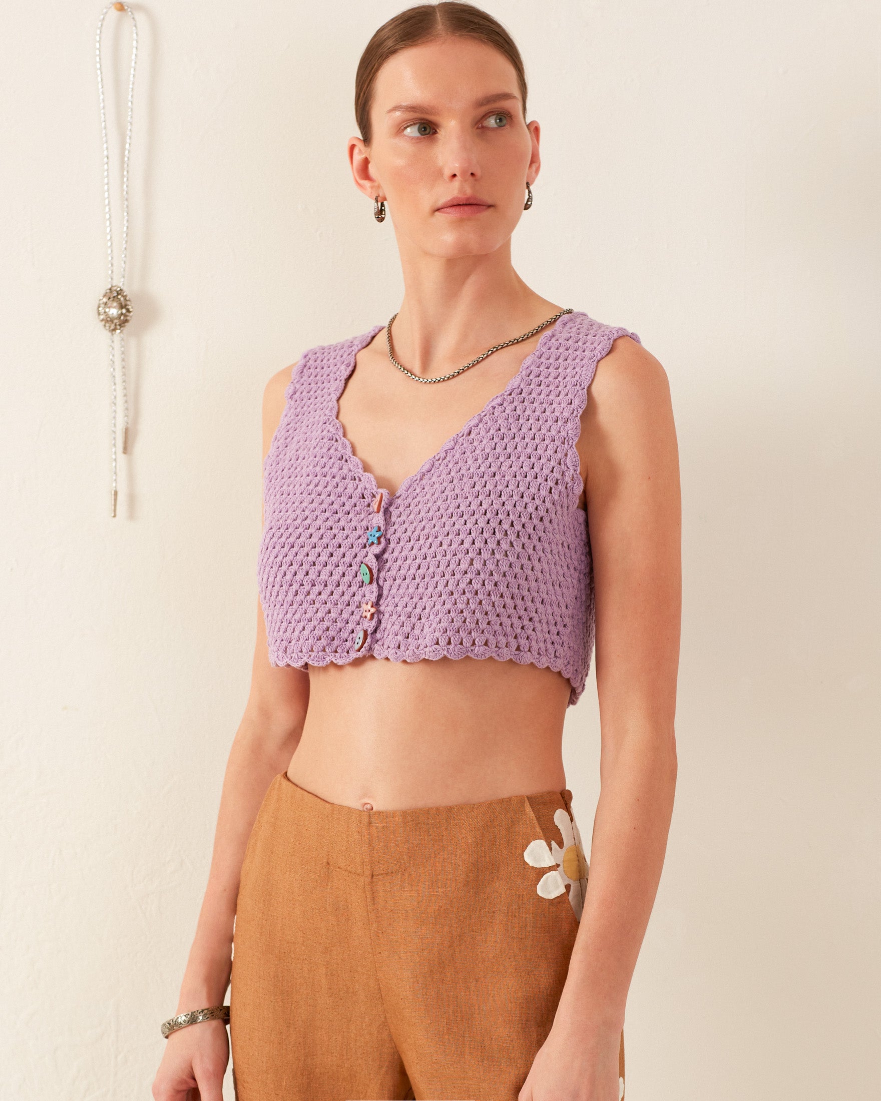 A person wearing the Ludi Lavender Crochet Top and brown pants adorned with white flower details stands against a light background. They have short hair and wear hoop earrings, necklaces, and bracelets.