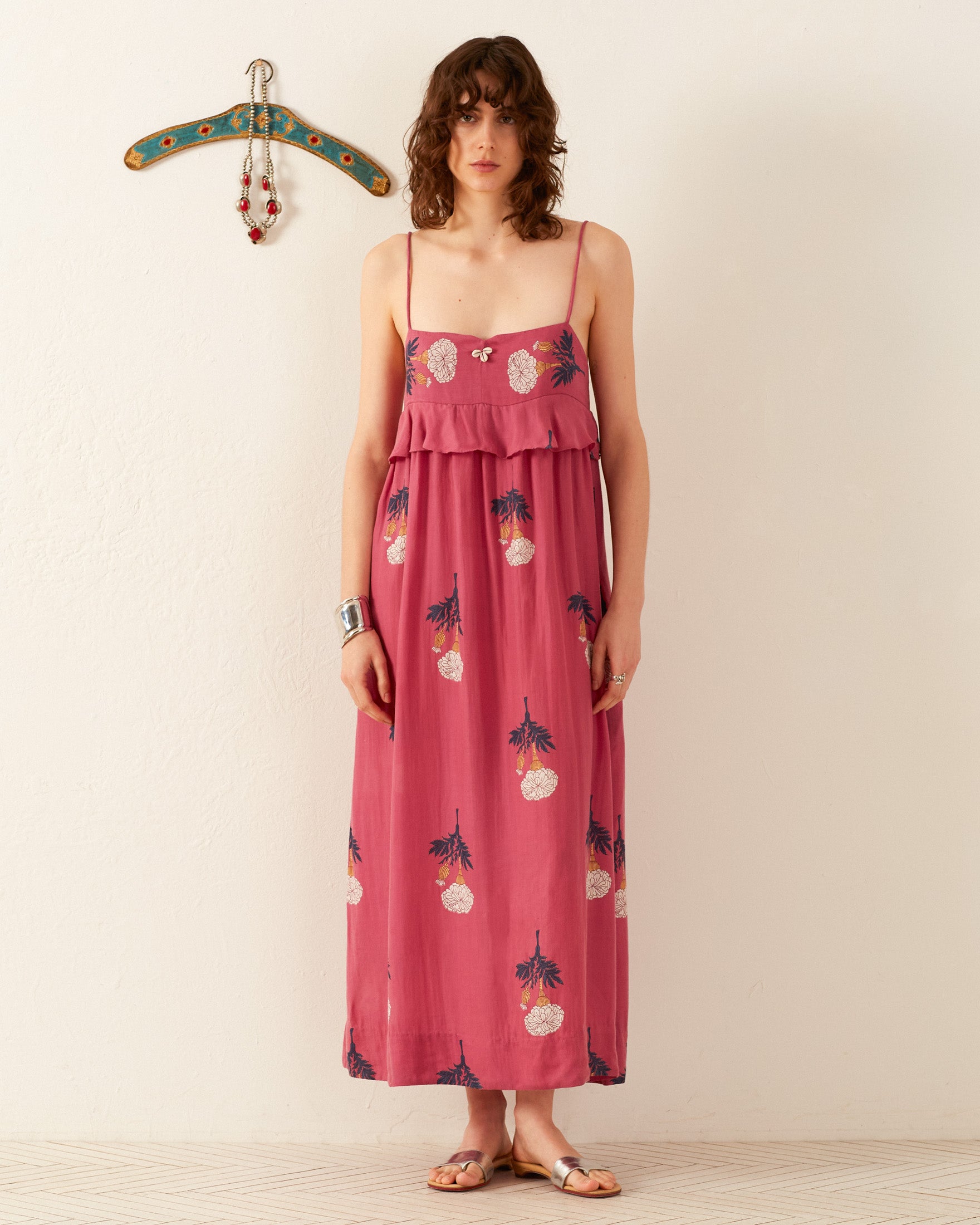 A person stands against a light wall, wearing the Martina Hibiscus Rose Dress, a flowing summer dress featuring a pink floral print and paired with sandals. Made from Tencel fabric, it adds an elegant touch to the ensemble. In the background, a decorative hanger displays jewelry mounted on the wall.