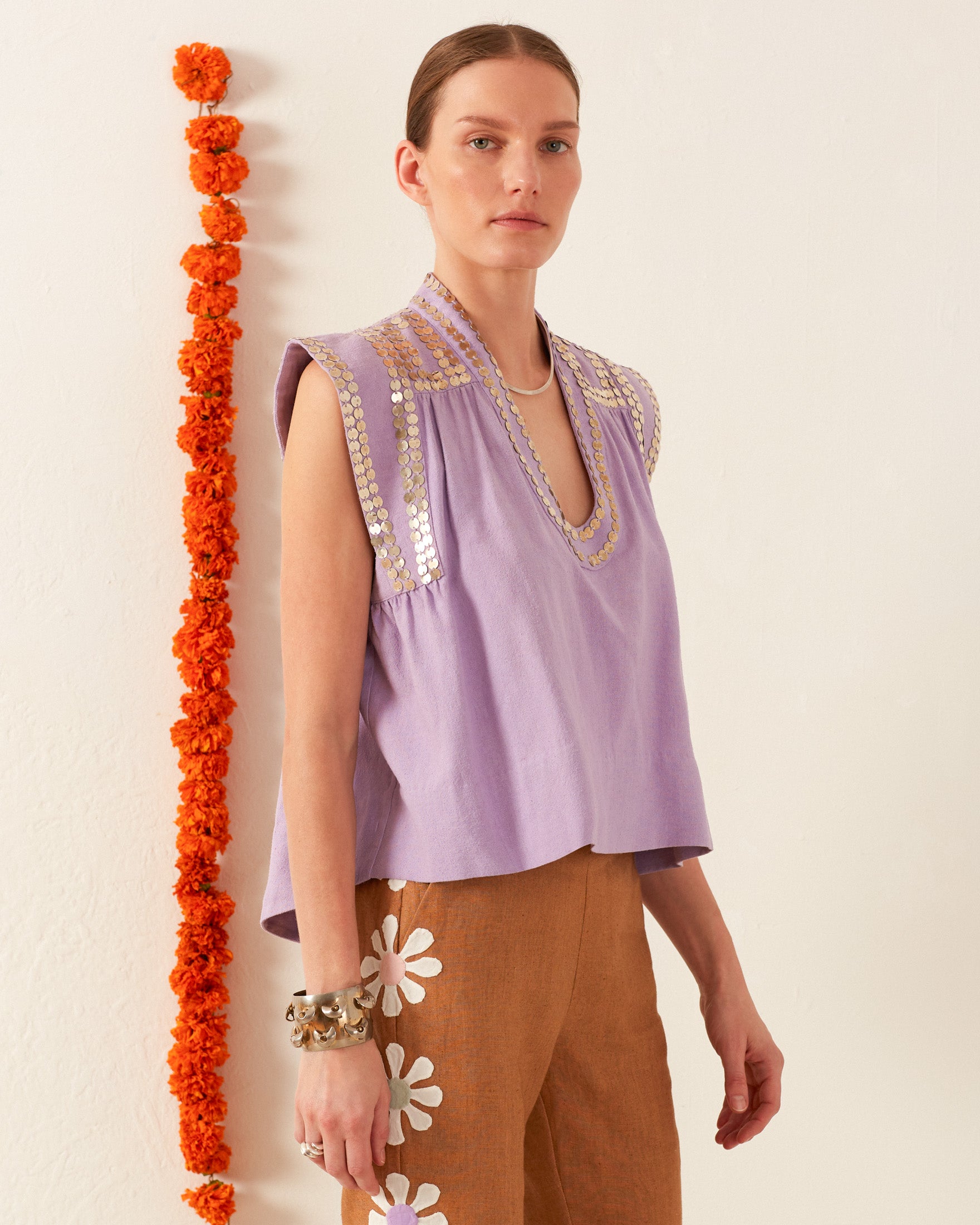 A person stands against a white wall wearing the Nehru Lavender Coin Top, paired with brown pants adorned with white floral designs. An orange garland and hand-stitched silver coins hang beside them, adding a touch of timeless elegance.