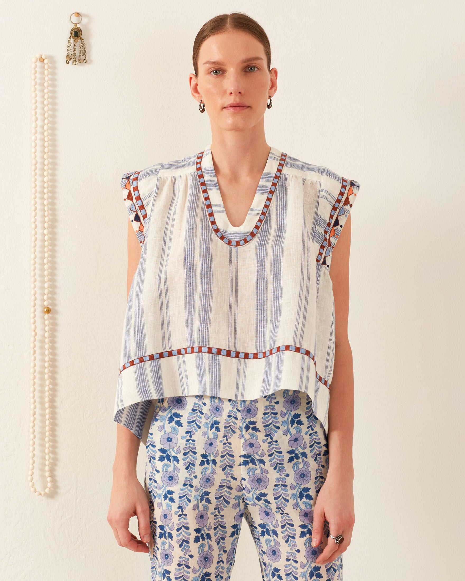 A person wearing the Nehru Pacific Embroidered Top, a vintage-inspired sleeveless blouse featuring blue and white stripes with patterned trim, stands against a plain wall. They pair it with floral-patterned pants. In the background, a string of pearls and a decorative tassel elegantly complement the ensemble.