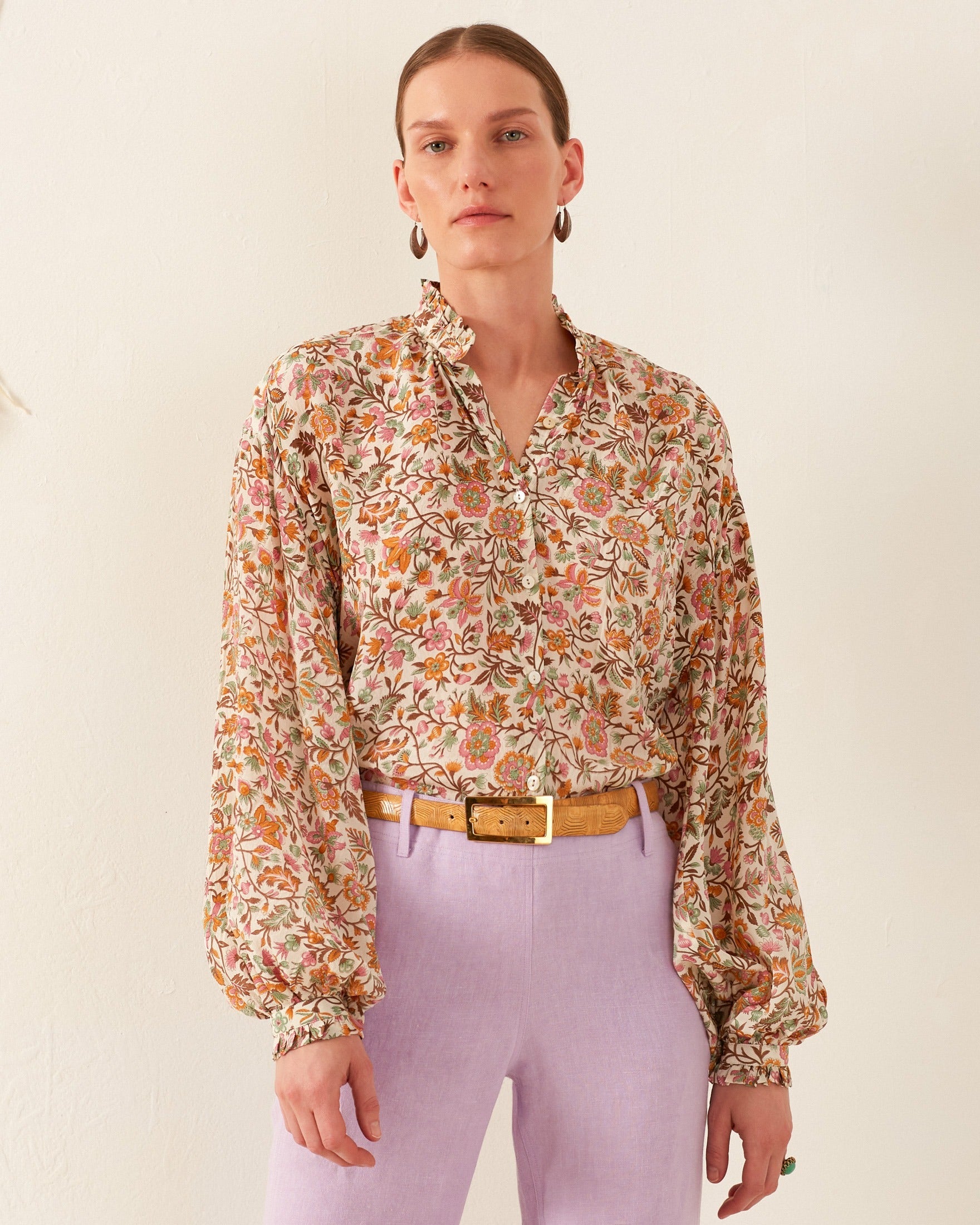 A person clad in the Poet Garden Party Silk Blouse, featuring a block-printed floral design with a ruffled collar and cuffs, stylishly paired with light purple pants and a tan belt. Their short hair enhances the ensemble as they stand against a plain background, gazing directly at the camera.