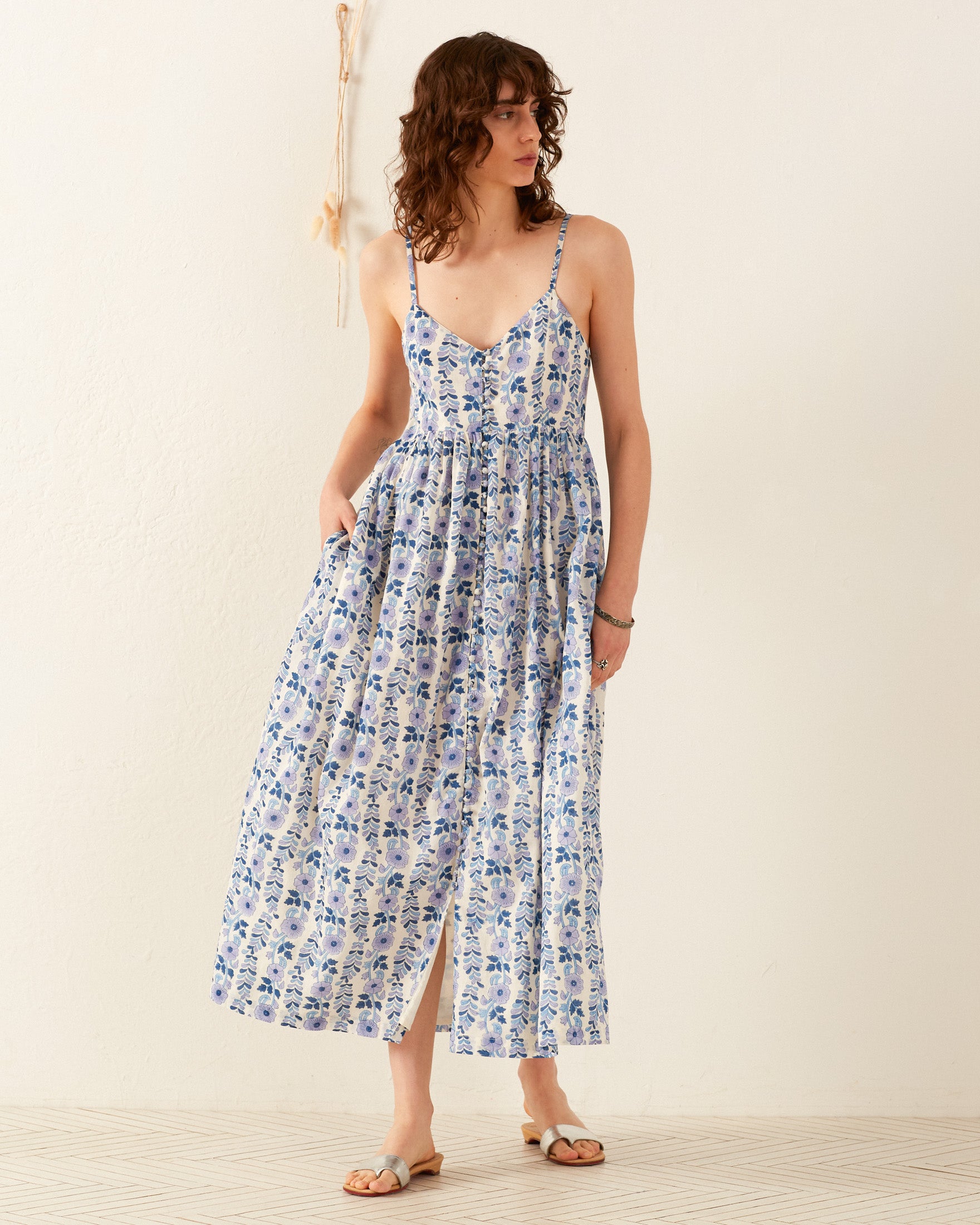 A person wearing the Skylar Iris Ivy Dress stands against a plain, light-colored background, looking to the side. The dress is a hand-block printed floral cotton voile maxi with spaghetti straps and features blue and white patterns. They are paired with light-colored sandals.