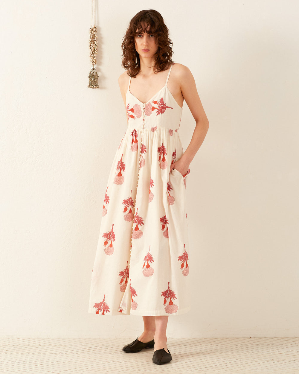 A person with shoulder-length hair stands against a light wall, wearing the Skylar Tea Rose Dress, a sleeveless summer outfit adorned with a red floral pattern. They sport black slip-on shoes and have one hand in a pocket. A decorative hanging is visible on the wall, adding charm to this vibrant scene.