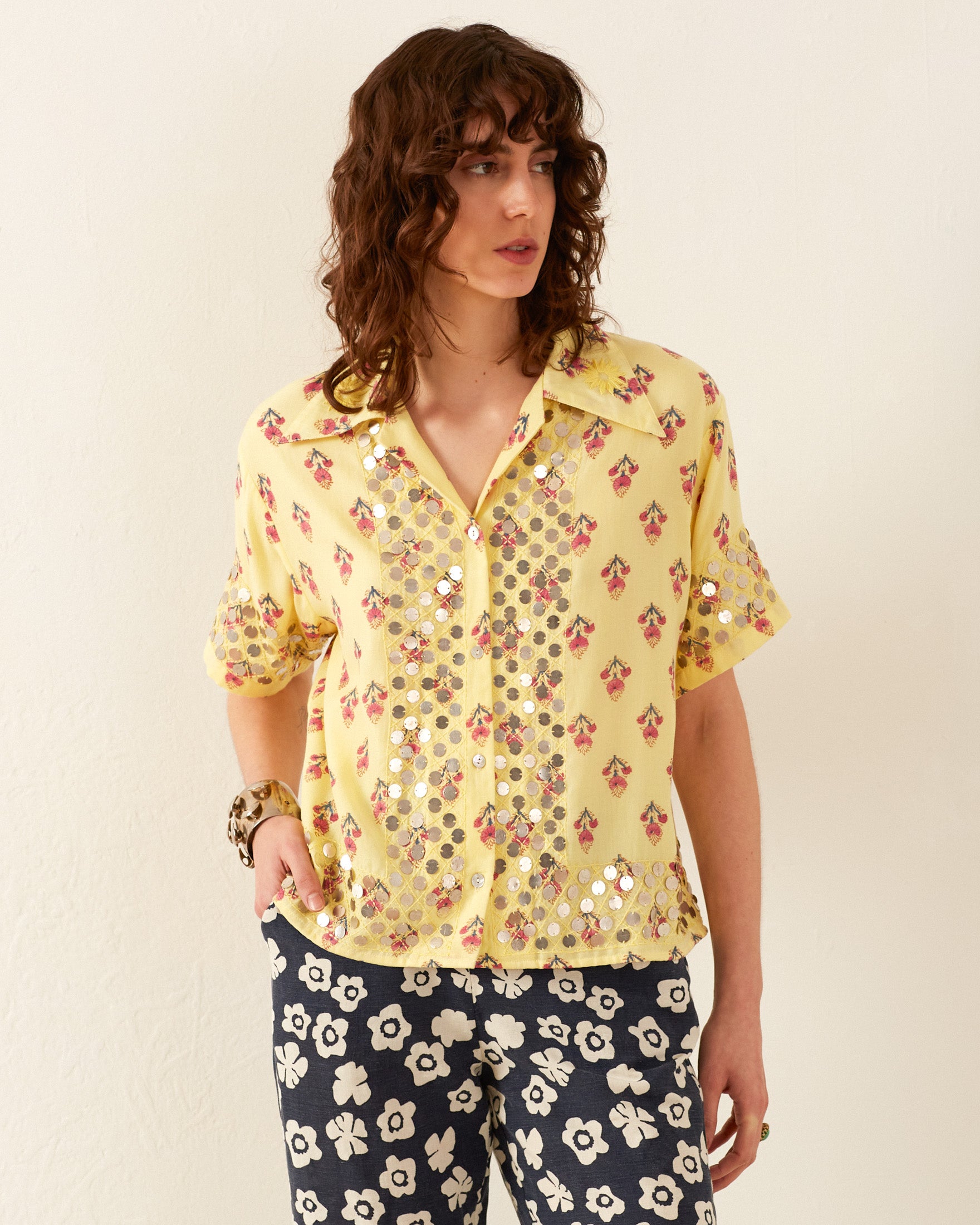 A person with wavy hair is wearing the Stevie Primrose Coin Shirt, a vintage-style Hawaiian shirt adorned with floral patterns and reflective sequins. They are paired with black pants featuring white flower designs, set against a minimal plain background.