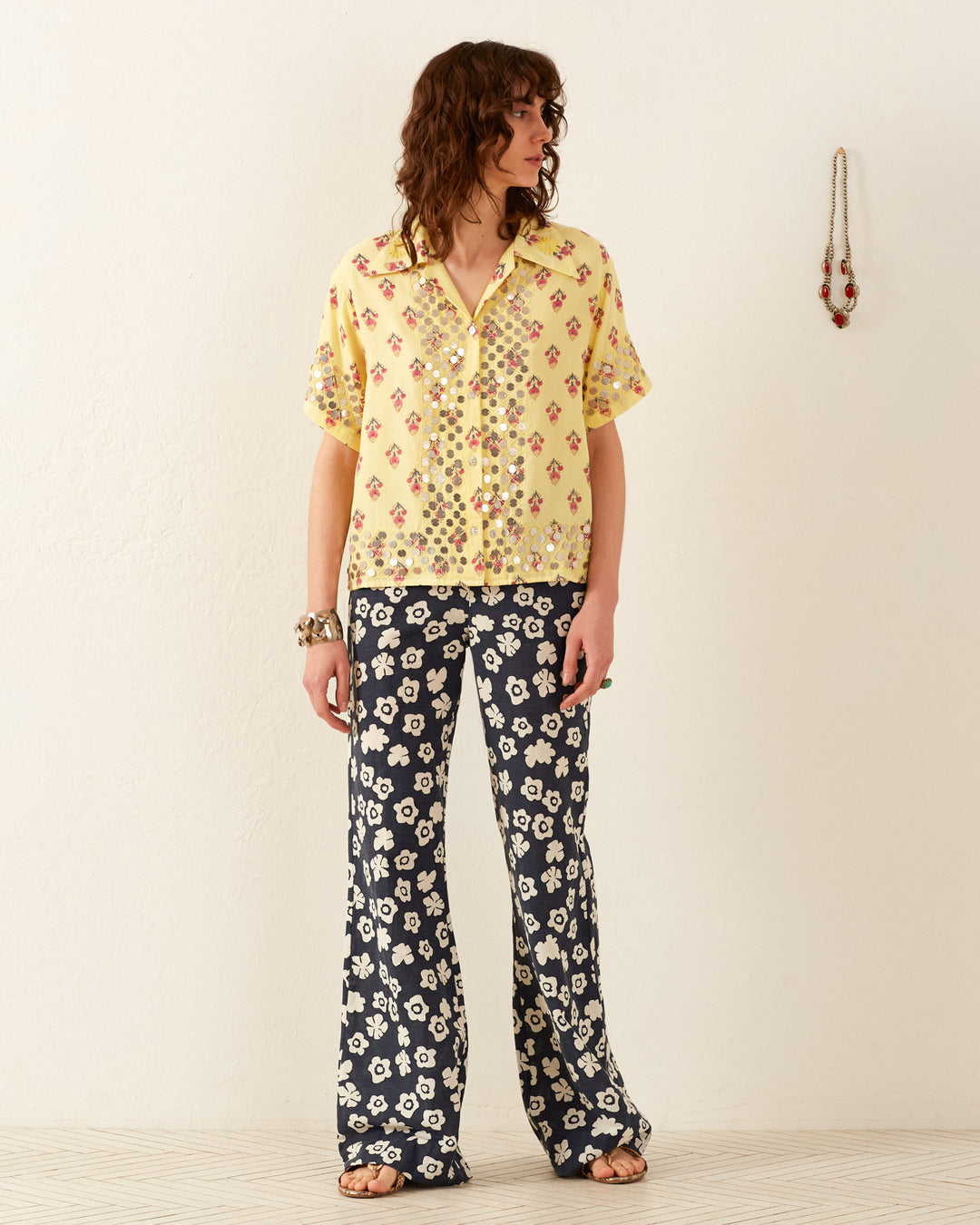 A person stands against a plain white wall, wearing the Stevie Primrose Coin Shirt, a vintage Hawaiian style with yellow short-sleeve floral designs. They pair it with black pants featuring white floral patterns, are barefoot, and there is a necklace hanging on the wall.