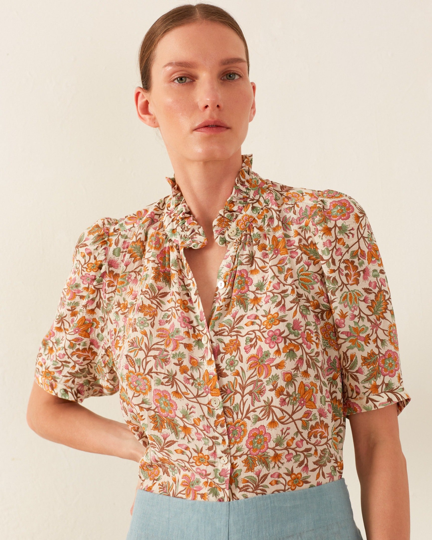 The person wears the Winn Garden Party Silk Shirt, a floral short-sleeved blouse featuring a ruffled collar, paired with light blue pants. This blouse displays a vibrant pattern of orange, pink, and green flowers against a plain, light-colored background.