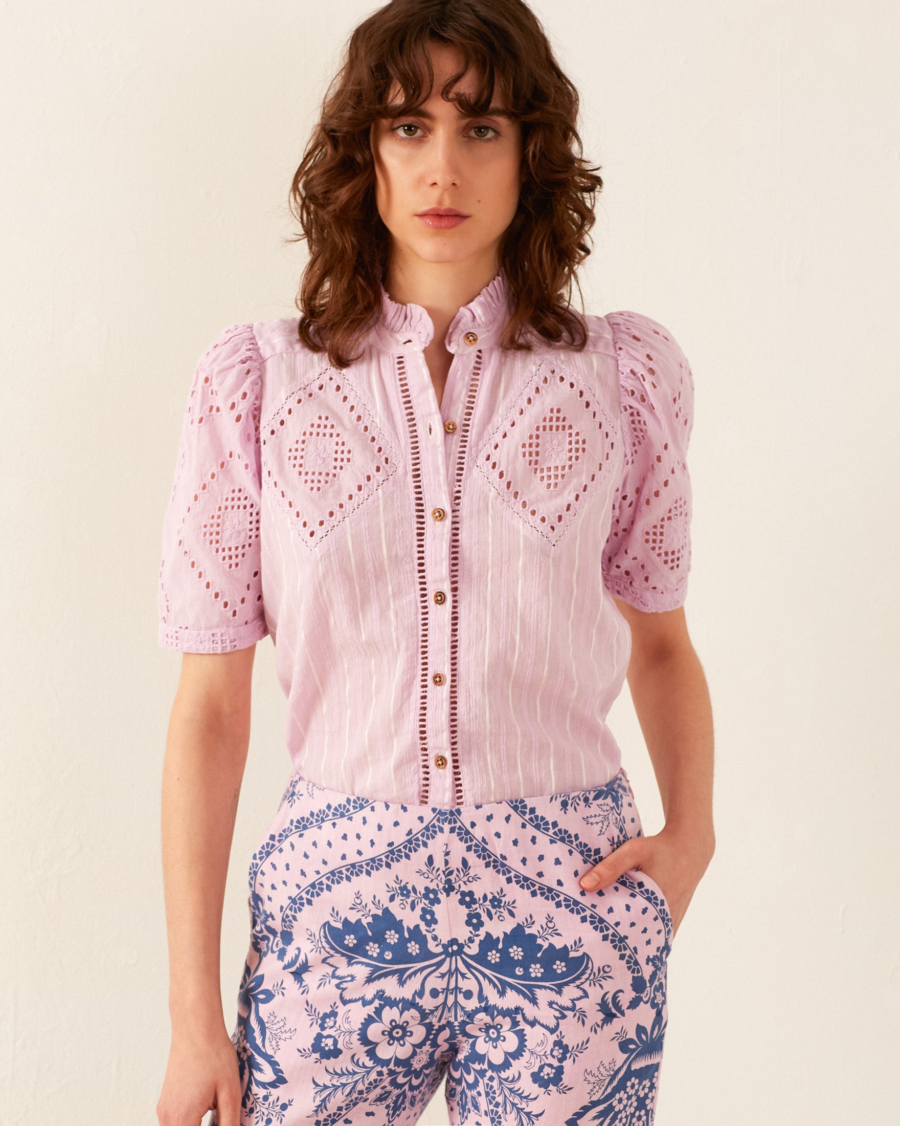 A person with shoulder-length wavy hair is wearing the Winn Lilac Eyelet Shirt, a light pink blouse showcasing hand-block printed geometric embroidery and puffed sleeves. They have paired it with '70s vintage-inspired blue and pink patterned pants while standing against a plain light background.