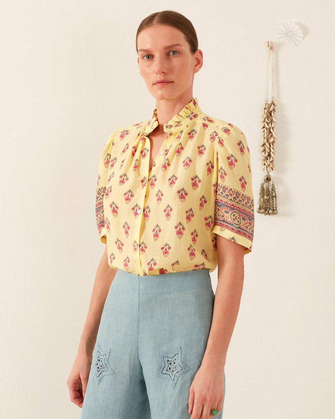 A person stands against a plain wall wearing the Winn Primrose Bhutti Shirt, which features a vintage-inspired floral motif, paired with blue pants adorned with star-shaped embroidery. The background is accentuated by a decorative hanging with shells and a bell.