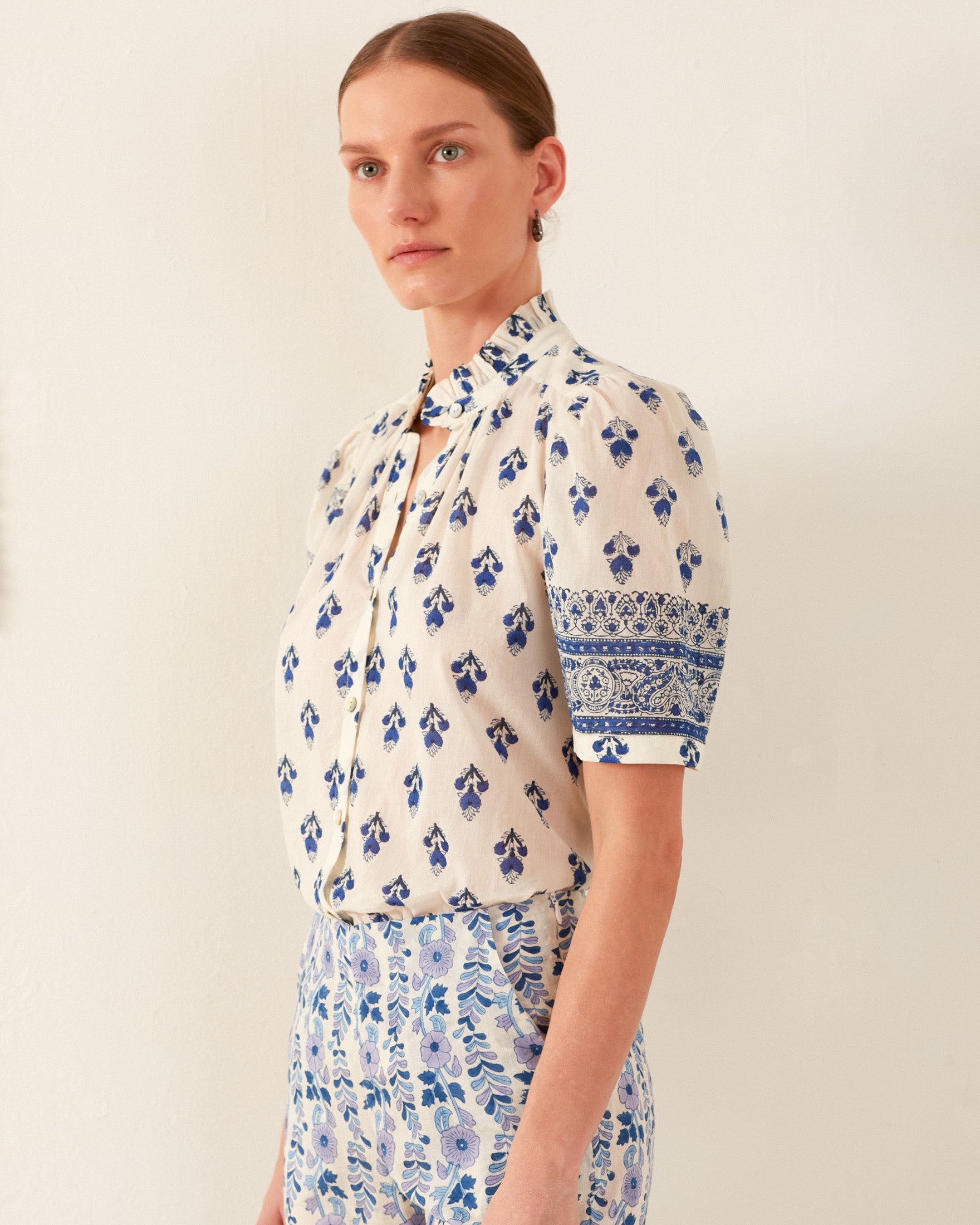 A person with their hair tied back is wearing the Winnie Indigo Bhutti Shirt, featuring a vintage-inspired blue and white floral design with short sleeves, paired with a skirt that boasts a similar intricate pattern. They are standing against a plain light-colored wall.