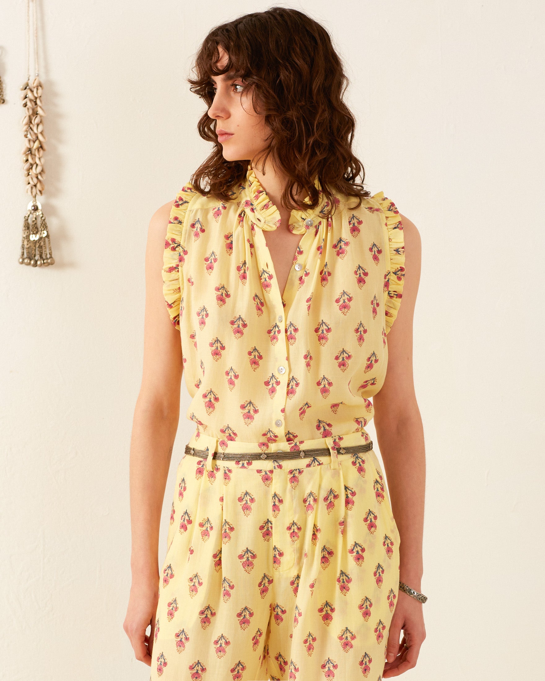 A person with wavy brown hair is wearing the Wylie Primrose Bhutti Shirt, a vintage-inspired sleeveless blouse in yellow with a floral pattern, along with matching pants. They stand against a light-colored wall adorned with decorative hanging items in the background.