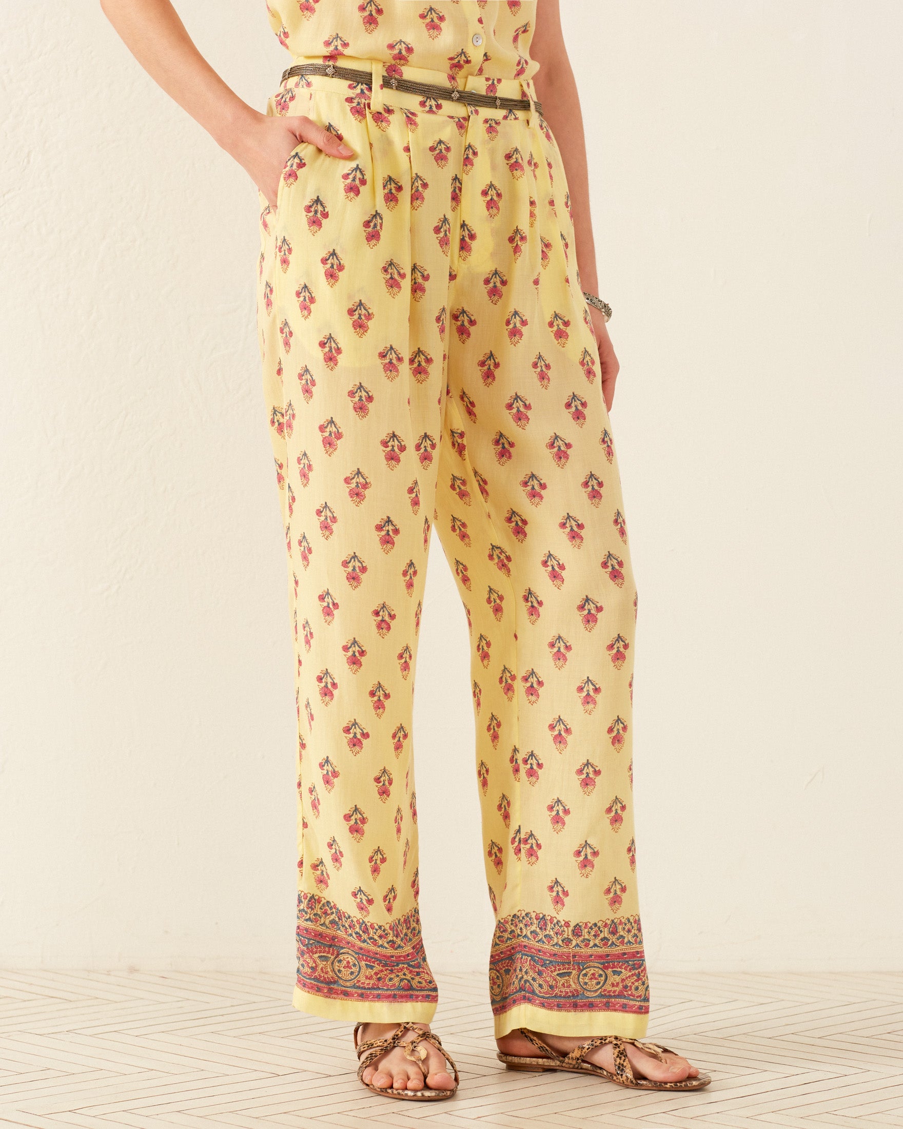 A person wearing the Colette Primrose Bhutti Pant, characterized by its high-waisted design, yellow pattern, lace panels, and accessorized with a brown belt and sandals, stands on a tile floor.