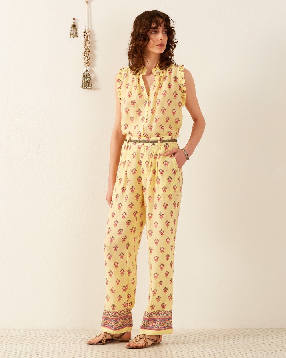 A person stands against a cream wall, dressed in the Colette Primrose Bhutti Pant and a yellow patterned blouse. They are barefoot with curly hair, while a decorative tassel adorns the wall.