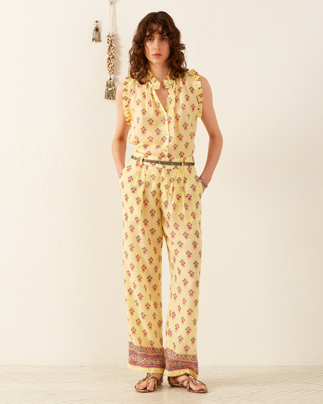 A person wearing the Colette Primrose Bhutti Pant stands against a white background, with tassels hanging on the wall, perfectly complementing the high-waisted design.