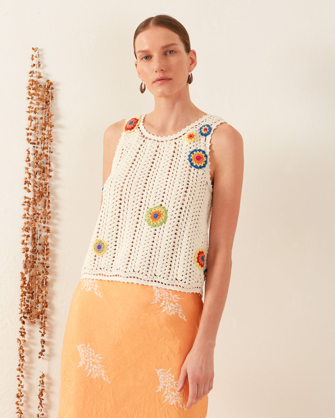 A person stands against a plain background wearing the Lula Pearl Flower Crochet Top, a vintage-inspired sleeveless top in white, featuring colorful floral patterns with a daisy motif. They pair it with a long orange skirt accented by white designs and accessorize with hoop earrings, while their short hair is styled slicked-back.