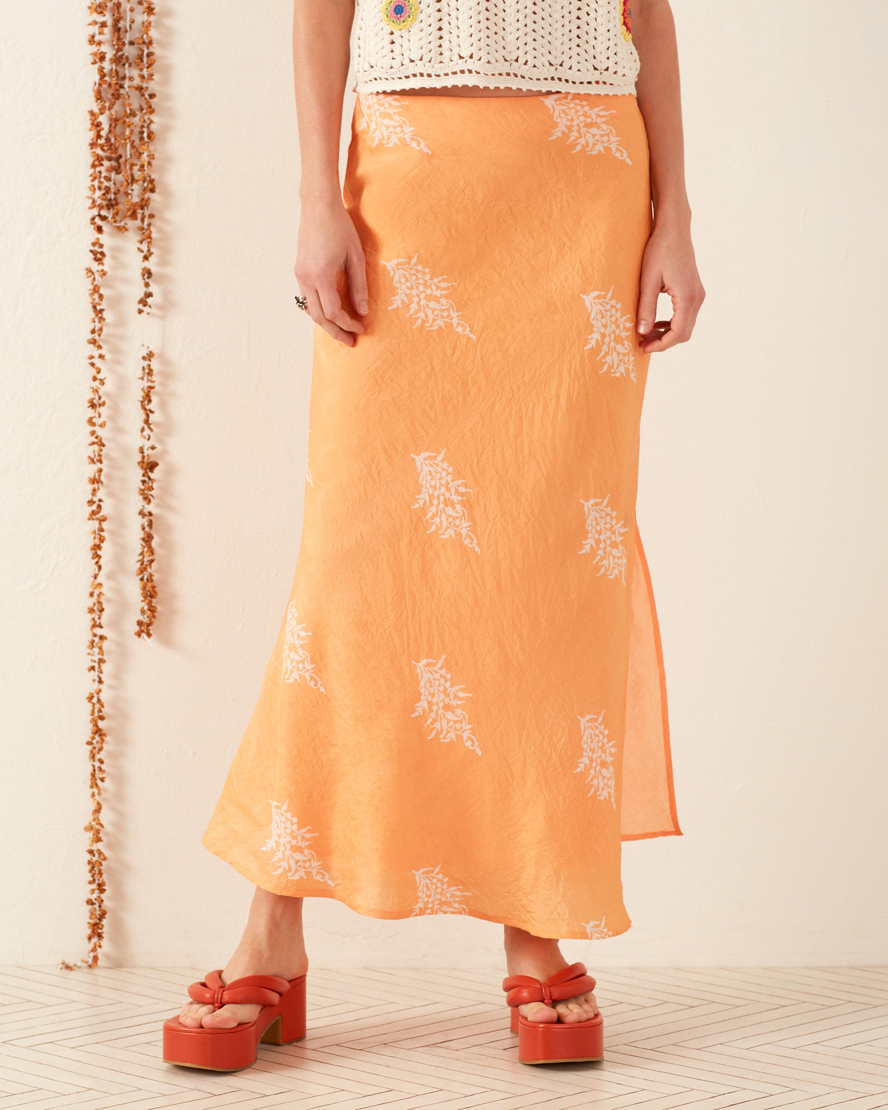 A person in a Lyra Hibiscus Rose Silk Skirt and platform sandals stands on a textured floor, flawlessly paired with an ivory cotton sleeveless tank adorned with floral appliqués.