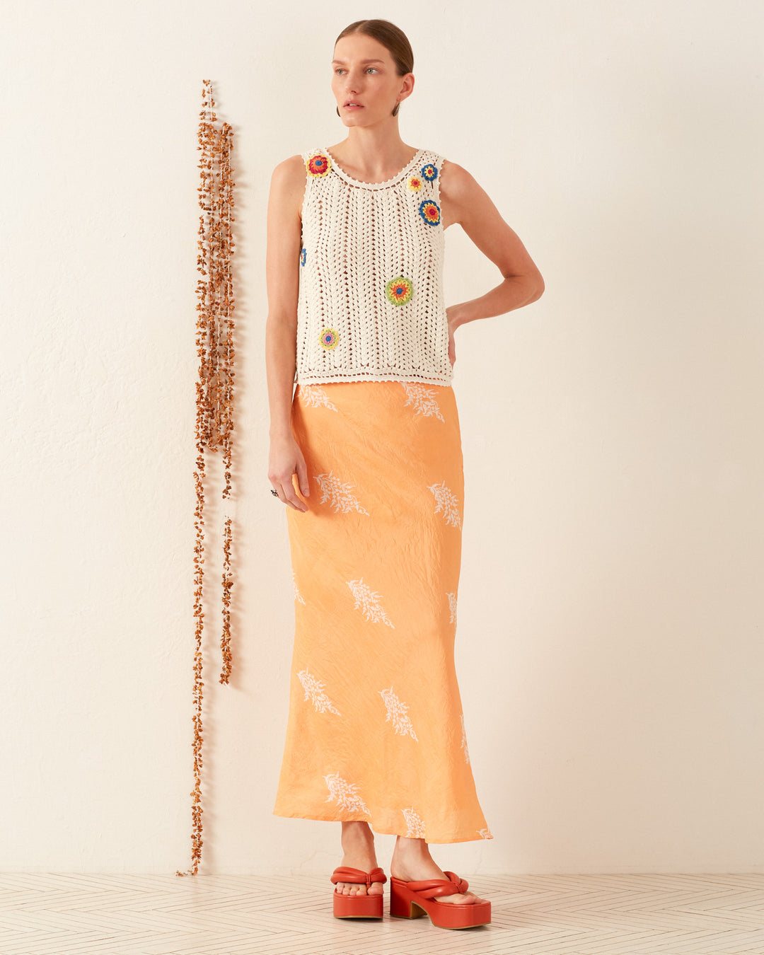 A person stands against a white wall wearing the Lyra Hibiscus Rose Silk Skirt, a white crochet top adorned with floral appliqués, and orange platform sandals. A string of small hanging objects is visible on the wall.