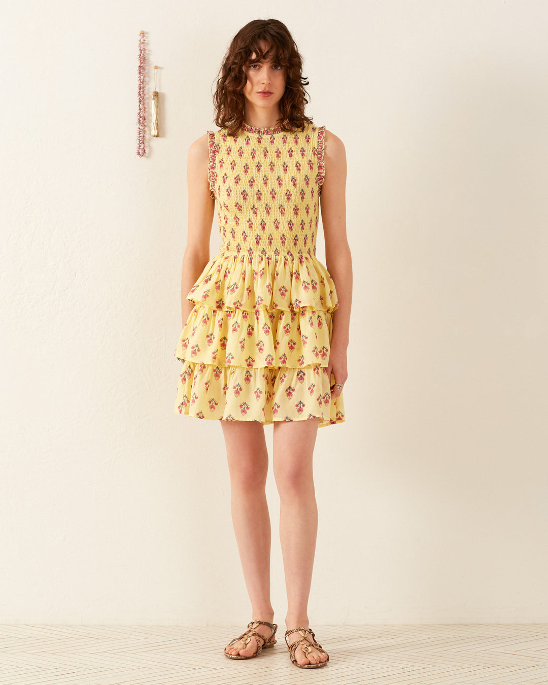 A person wearing the Caeli Primrose Bhutti Dress, featuring a sleeveless design and yellow floral patterns with tiered ruffles, stands against a plain white background, evoking a seaside feel.