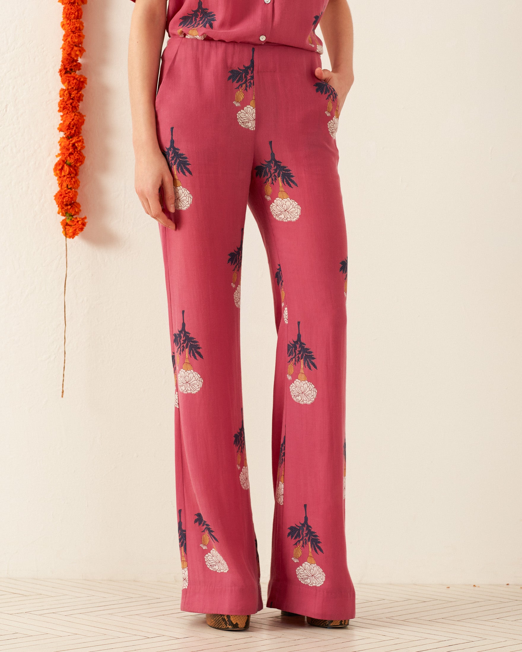 A person wearing the Charlie Hibiscus Rose Pant, featuring pink, retro floral and paisley designs in a wide-leg style with hands in pockets, stands beside an orange marigold garland hanging on a cream wall. The floor features a subtle geometric pattern.