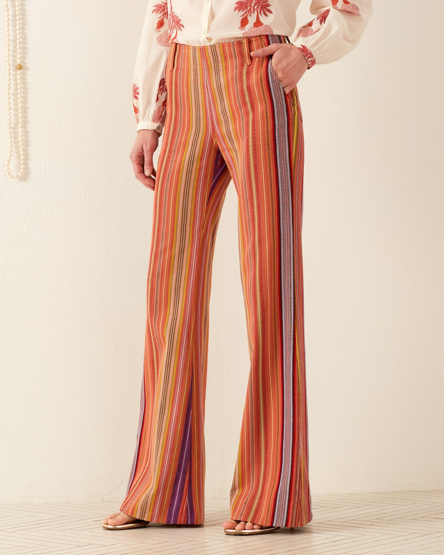 A person wearing the vibrant Charlie Melon Stripe Ikat Pant and a white blouse with red floral embroidery stands on a tiled floor against a cream-colored wall.