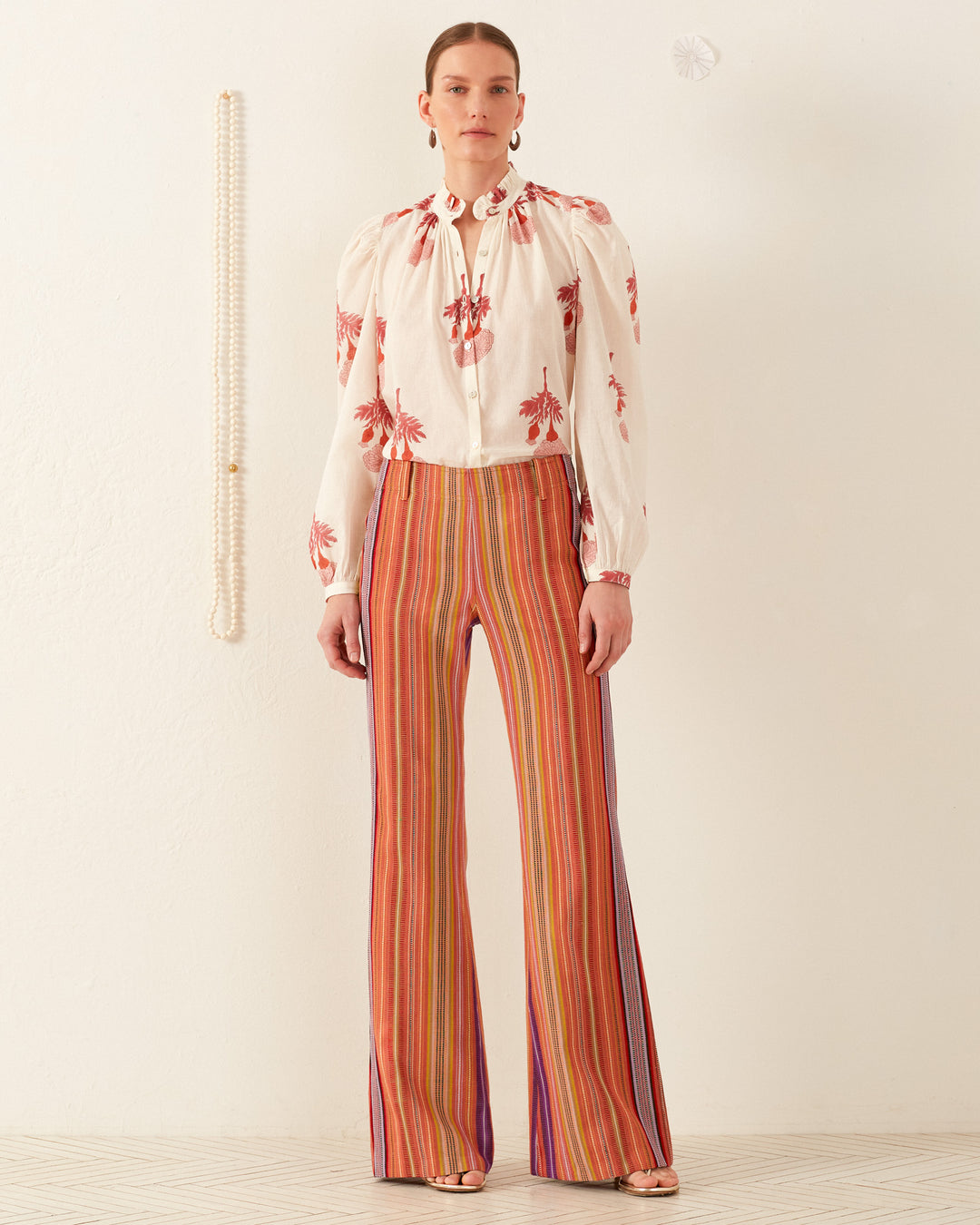 A person stands against a plain wall wearing a floral white blouse and the Charlie Melon Stripe Ikat Pant, showcasing vibrant '70s-style flair. The blouse features red floral patterns and puffed sleeves. The patterned floor adds charm, while a strand of pearls elegantly hangs on the wall, completing the retro vibe.