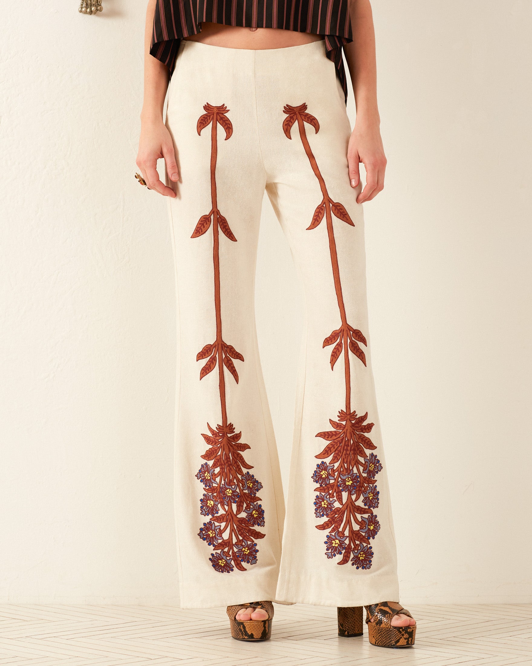 The person is wearing the Charlie Waterlily Bloom Pant, which boasts a vintage cream color and features intricate red floral embroidery cascading down the legs. They stand indoors on a subtly patterned floor, accented with brown platform heels. Their dark top showcases a chic striped pattern that perfectly complements the embroidered elegance of their pants.