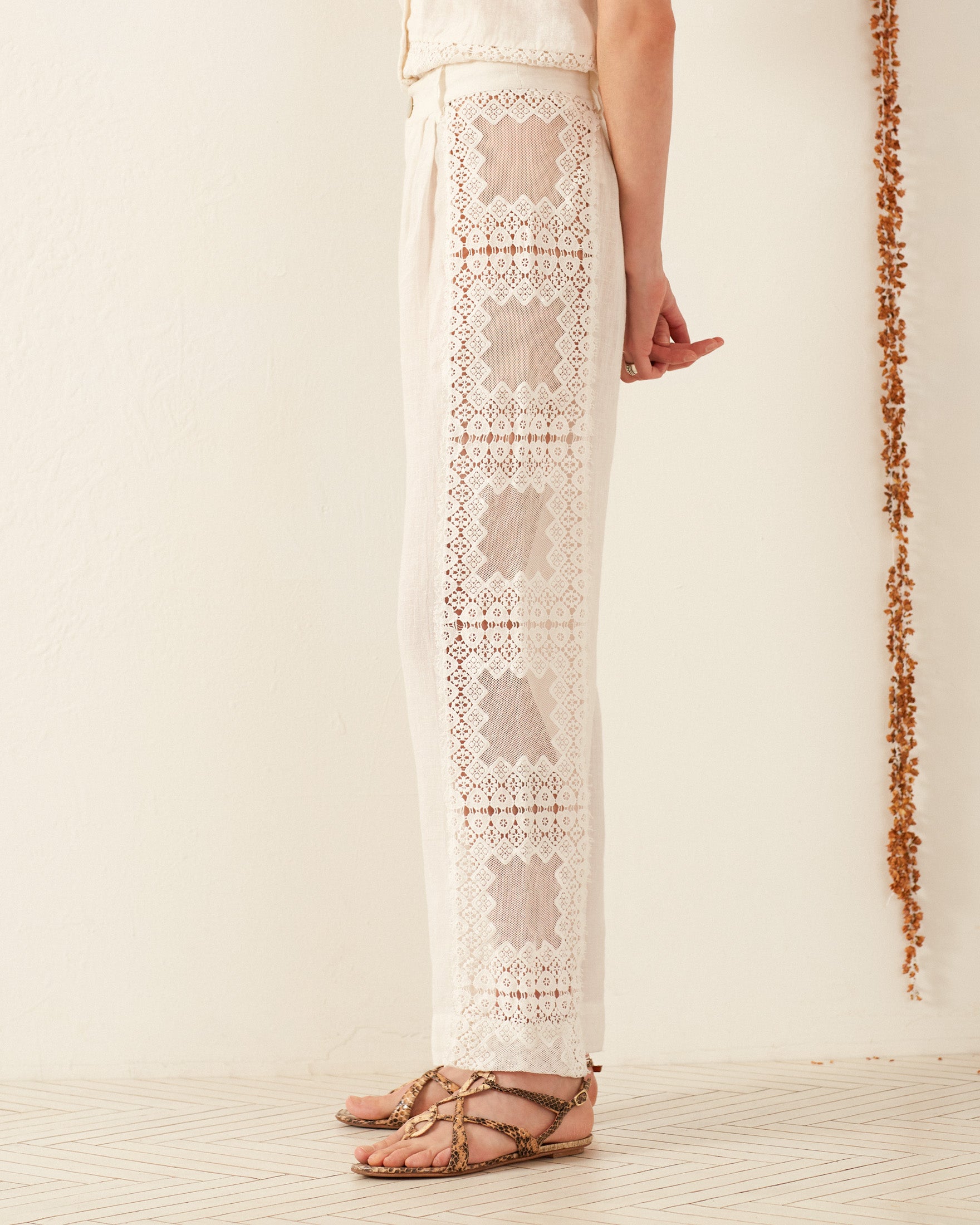 A person stands in profile wearing the Colette Picnic Eyelet Pant, featuring a lilac hue and intricate lace patterns along the sides. They are barefoot on a white tiled floor, with a brown tassel decoration adorning the white wall in the background.