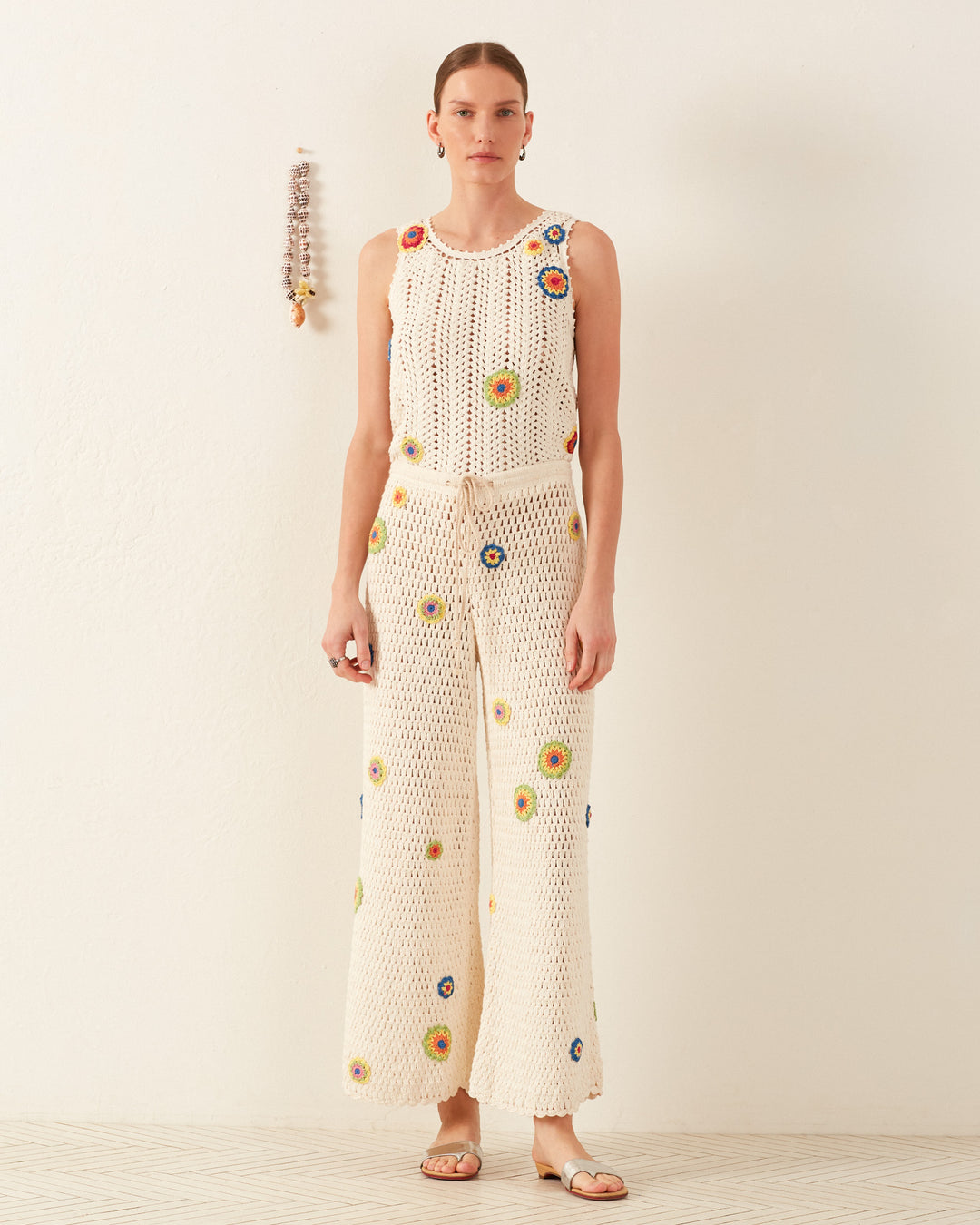 A person stands against a plain wall, dressed in the Elia Pearl Flower Crochet Pant set featuring a sleeveless, cream-colored crochet top and matching wide-leg pants with vibrant floral designs. They have short hair and are wearing flat sandals that beautifully enhance the sophisticated silk chiffon look of the ensemble.