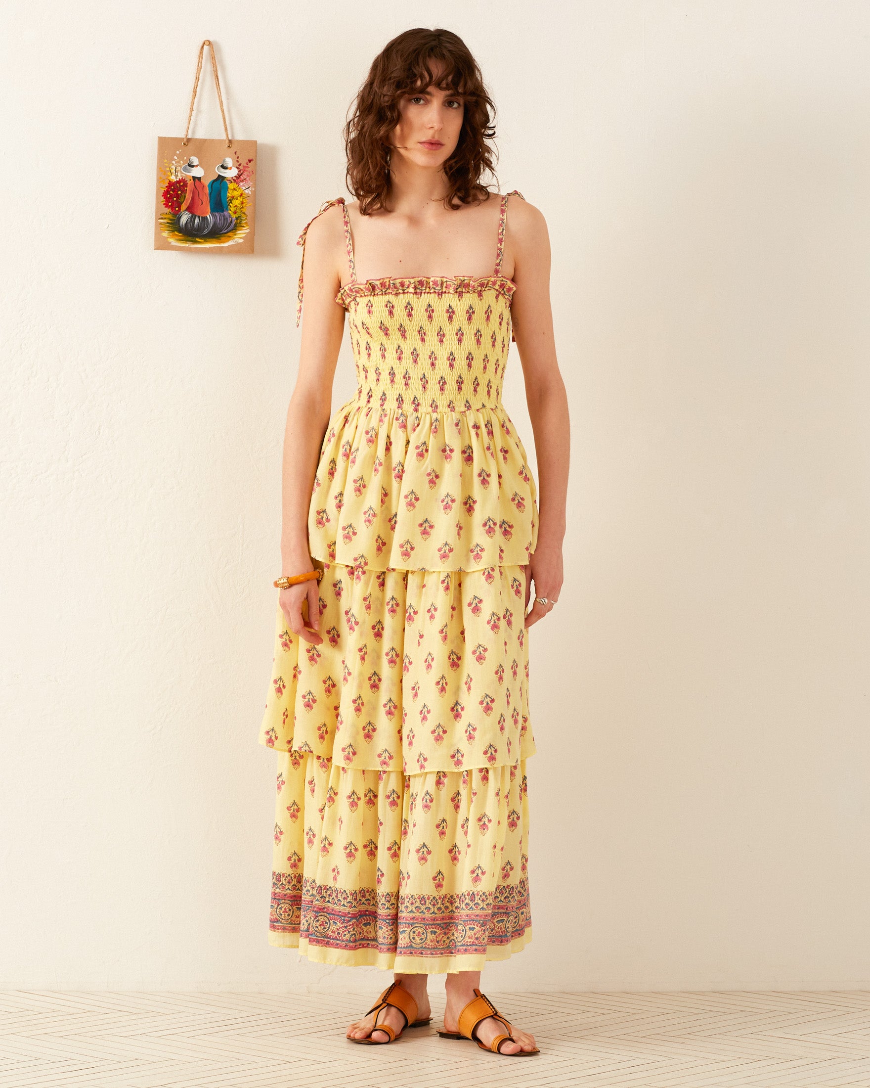 A person wearing the Kaia Primrose Tier Dress made of yellow floral cotton voile stands against a white wall. They have curly hair and are wearing sandals. A colorful painting hangs on the wall in the background, complementing their vibrant attire.