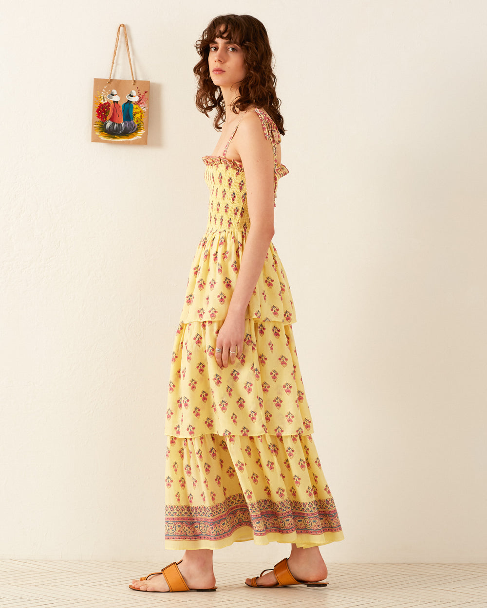 A person with curly hair stands sideways wearing the Kaia Primrose Tier Dress, a layered yellow cotton voile creation, featuring brown patterns at the hem. In a room with a white wall and a small colorful painting hanging on it, they complete their look with brown sandals.