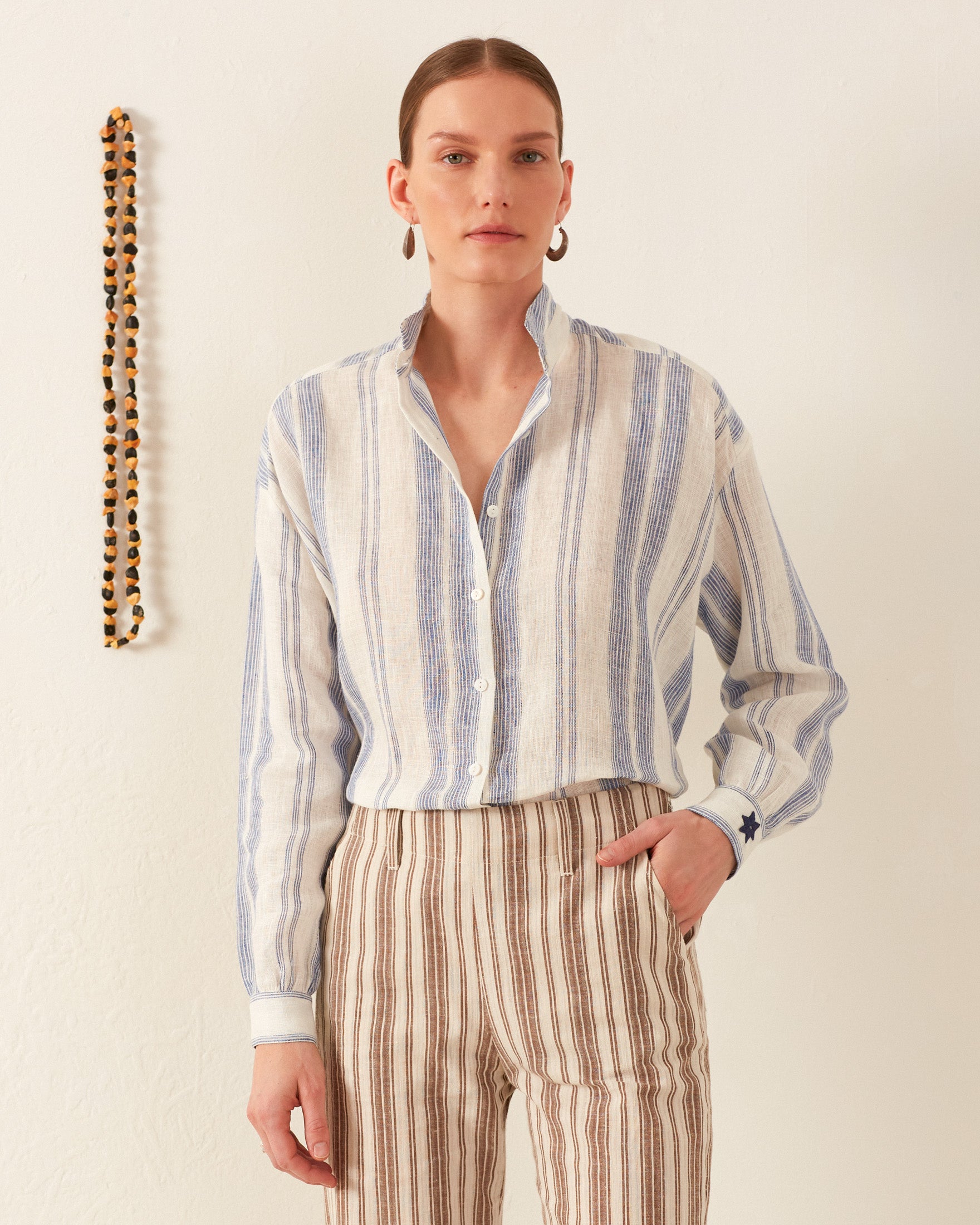 A person wearing the Kiki Pacific Star Shirt, featuring a hand-block printed blue-and-white floral design, along with striped pants stands against a plain wall. A string of beads hangs on the left side. They have a neutral expression and short hair.