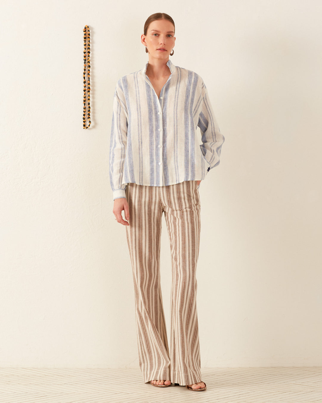 A person stands against a plain wall, wearing the Kiki Pacific Star Shirt, a blue-and-white floral print top, paired with brown and white striped pants. A string of beads hangs on the left. The floor is white with a subtle pattern, creating a serene backdrop for the hand-block printed cotton voile ensemble.