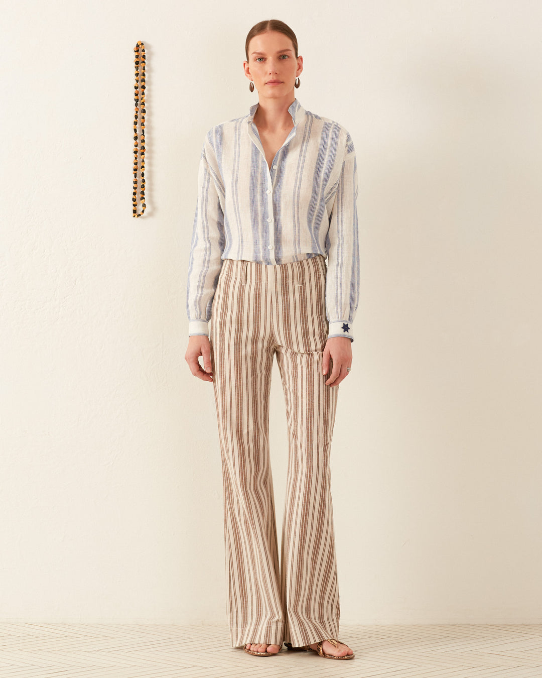 Against a light background, an individual wears the Kiki Pacific Star Shirt, which is hand-block printed and features vertical stripes in light tones. They pair it with wide-leg pants made from cotton voile. A decorative beaded string hangs on the wall beside them.