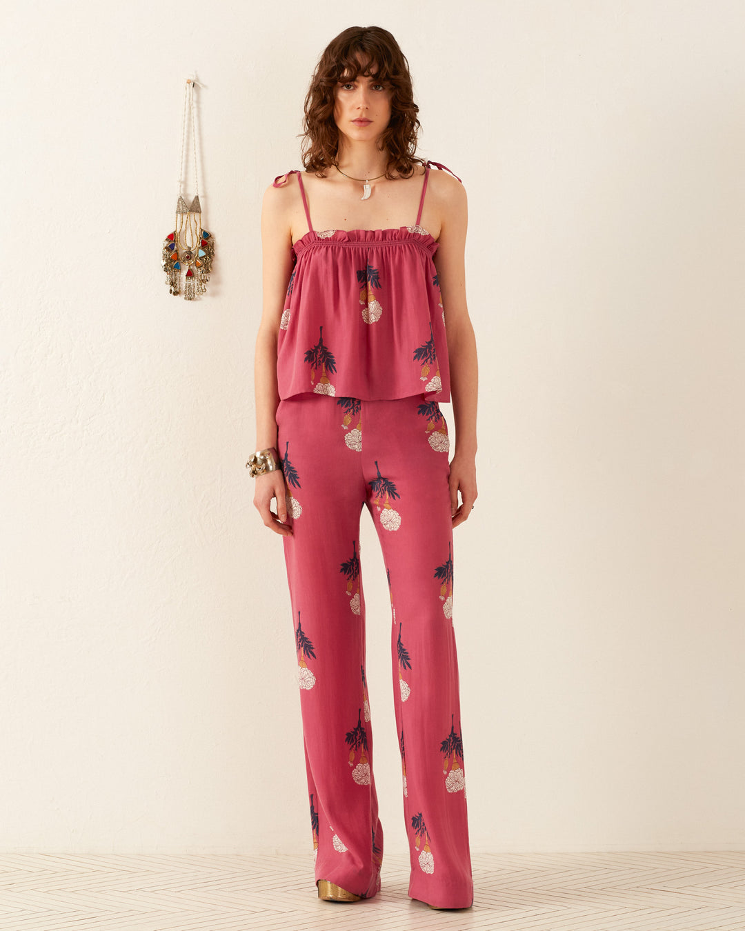 A person in a Lila Hibiscus Rose Top and matching wide-leg pants featuring a playful pineapple pattern stands on a light floor against a beige wall. Nearby, colorful tassels add charm to the vintage-inspired decor.