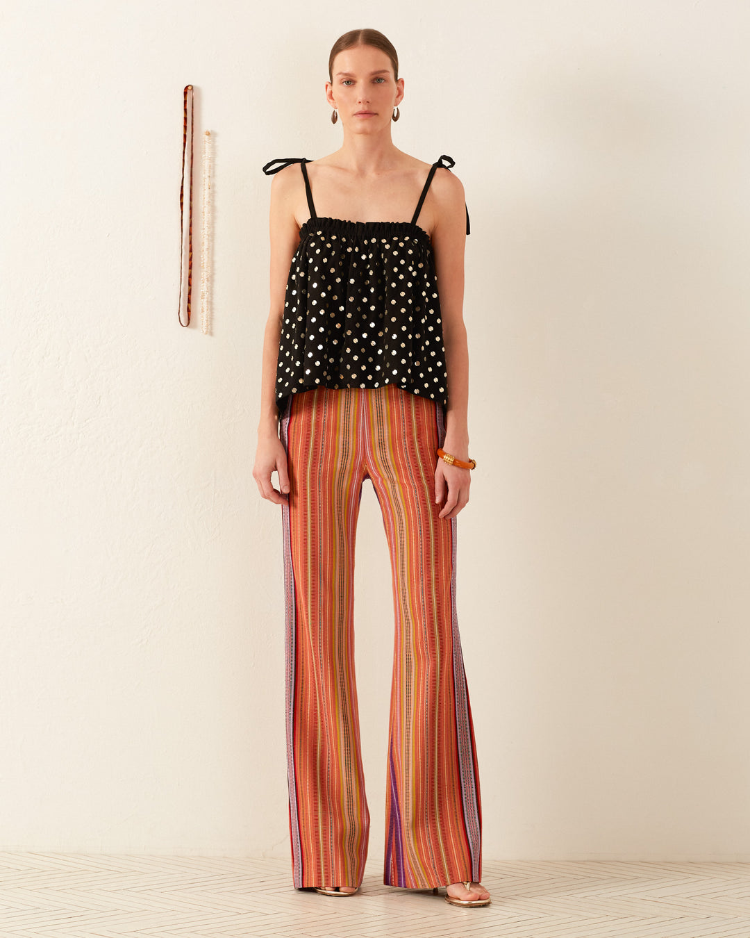 A person stands against a plain wall, wearing the Lila Night Sky Coin Top, a black hand-block printed sleeveless design with thin straps, paired with multicolored striped wide-leg pants. They have short hair, and there's a tape measure hanging on the wall.