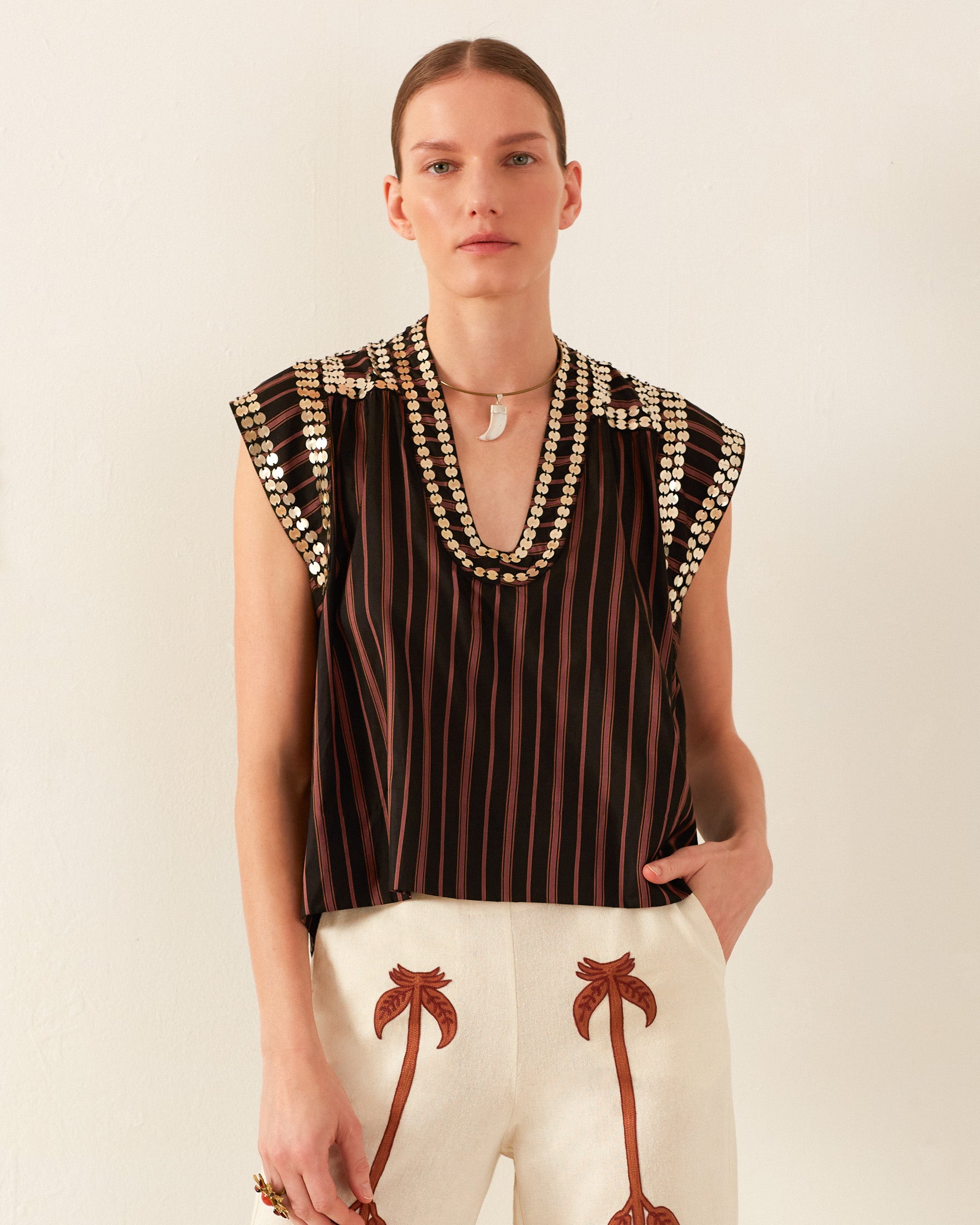 A person wearing the Nehru Clove Stripe Coin Top, which features Guatemalan ikat details and wood-carved buttons around the neckline, paired with white pants adorned with palm tree designs, stands against a plain background.