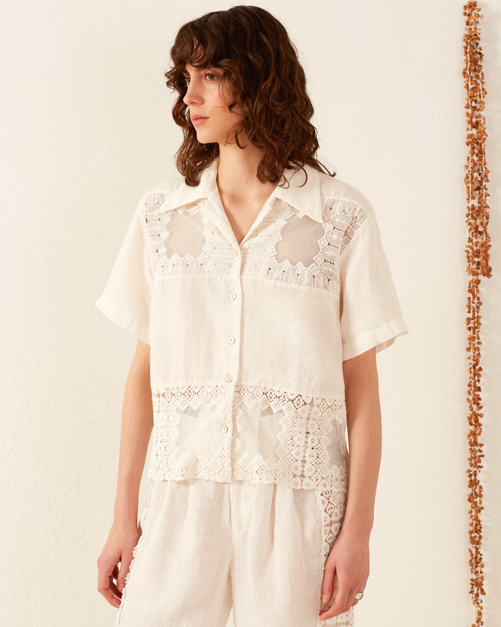 A person with curly brown hair wears the Stevie Picnic Eyelet Shirt, a white short-sleeve blouse featuring intricate lace patterns, paired with matching hand-loomed cotton pants. They stand against an off-white wall adorned by a decorative string of small objects hanging vertically.