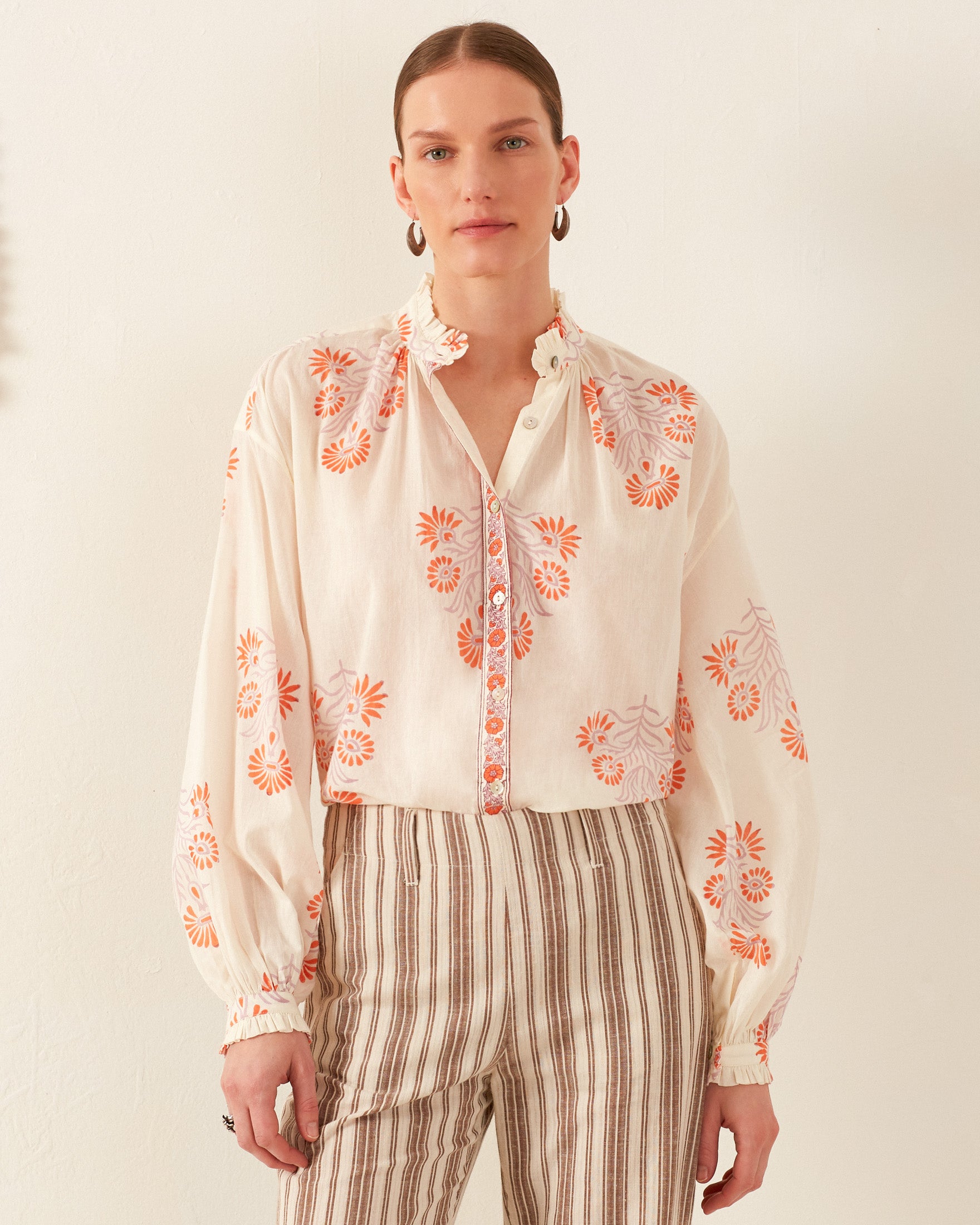 A person stands against a plain background wearing the Poet Papaya Aster Blouse, featuring a vintage-inspired design with orange floral patterns. They pair it with striped pants and have long hair tied back, accessorized with hoop earrings. The blouse's high collar adorned with ruffles adds an elegant touch to the ensemble.