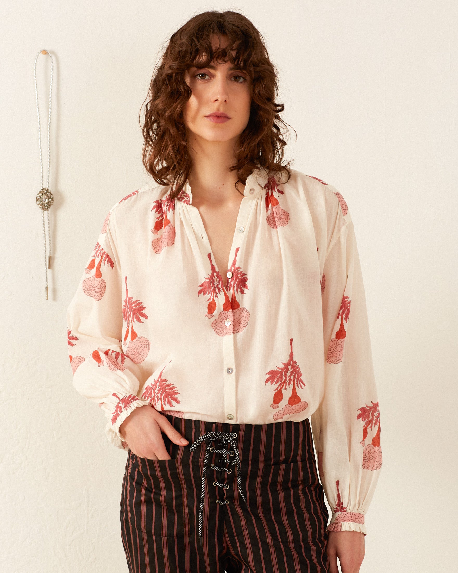 A person with long, curly hair wears the Poet Tea Rose Blouse, featuring a vintage-inspired design with red floral patterns, paired with dark pinstripe pants. They stand against a light-colored wall beside a decorative pendant.