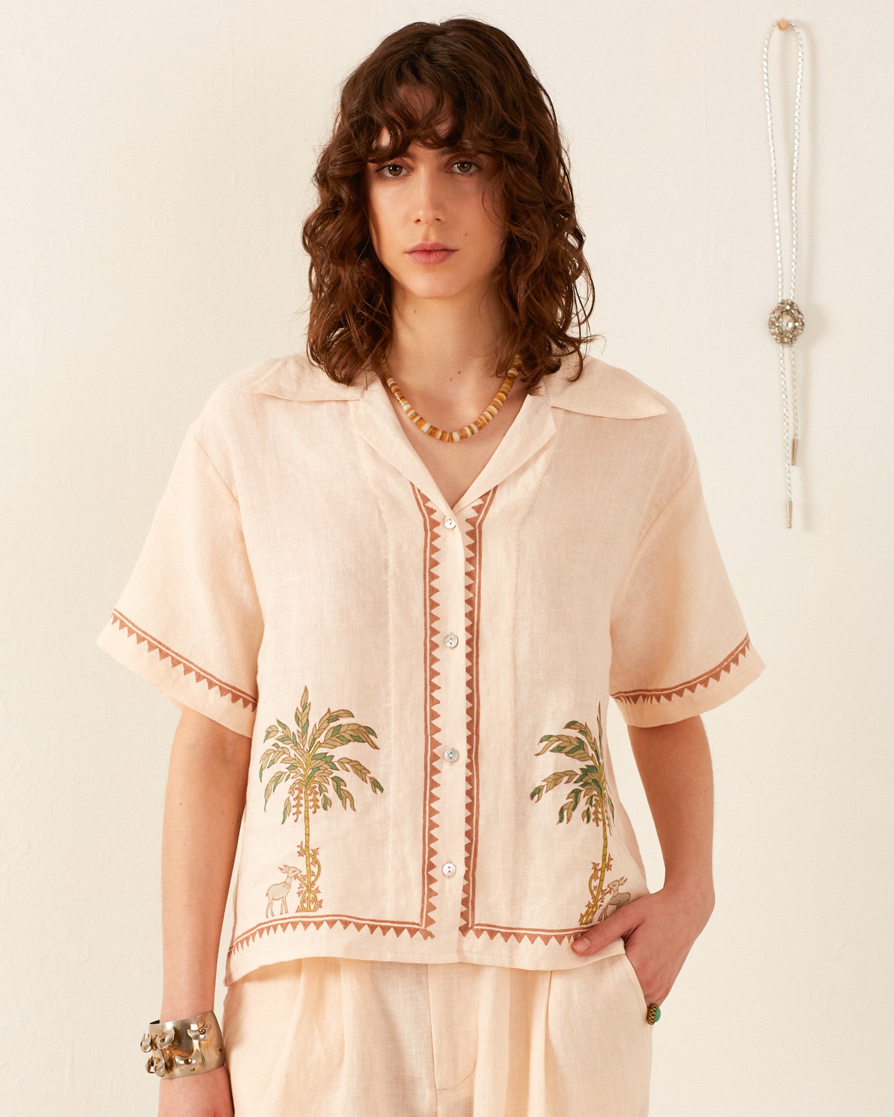 A person with wavy brown hair is wearing a Stevie Sanded Palm Shirt, which boasts vintage-style decorative palm tree embroidery. The background is a plain light wall adorned with a hanging ornament. The person has a neutral expression and complements their look with beaded jewelry.