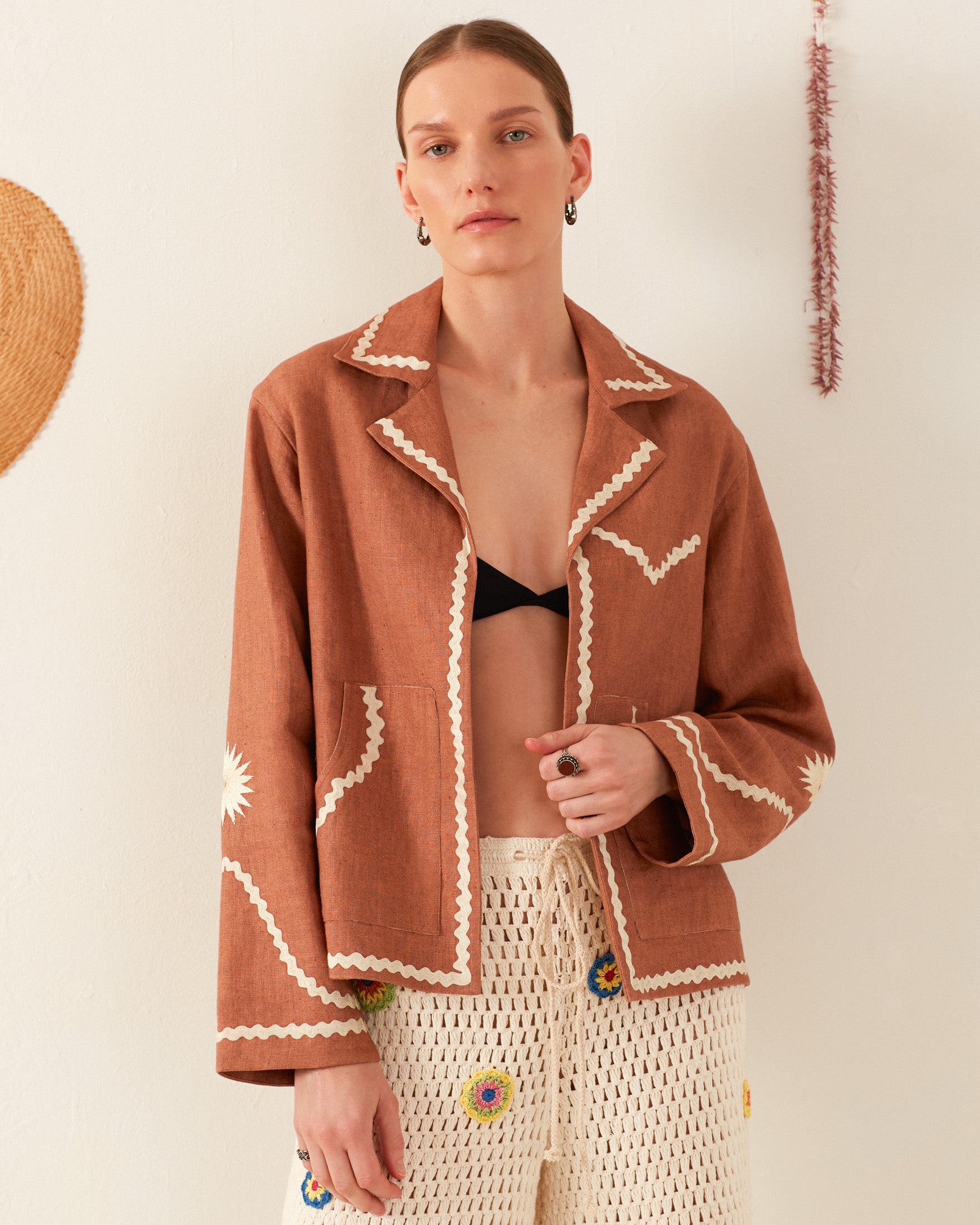 A person wearing a Western Clove Ric Rac Jacket over a black top and Charlie Deep Sea Daisy Pant stands against a light background. A retro floral cotton waistcoat pairs with earrings and a ring, while bright decorations on the wall enhance the lively aesthetic.