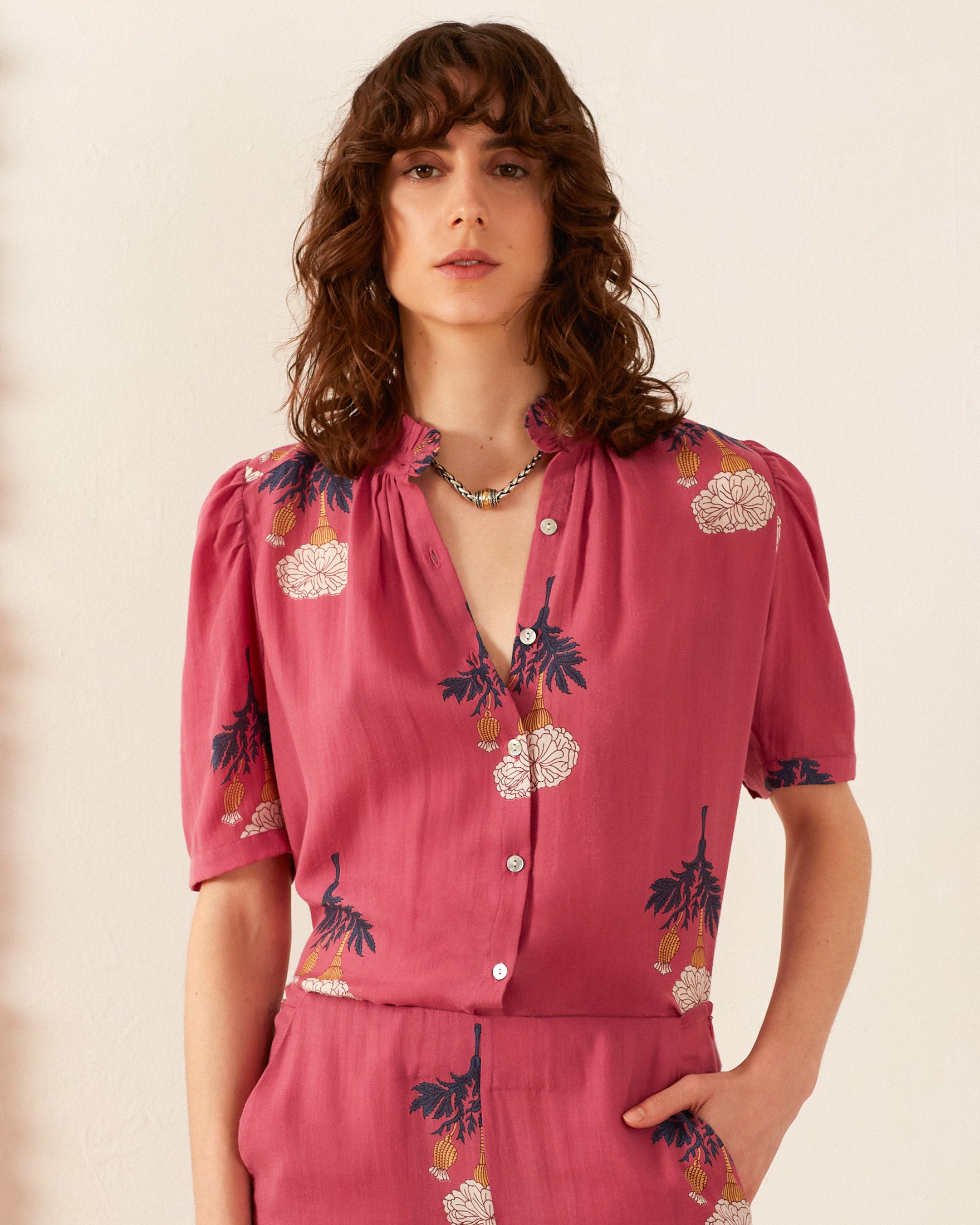 A person with wavy brown hair is wearing the Winn Hibiscus Rose Shirt, a vintage-inspired pink blouse with floral patterns, and matching pants. They stand against a plain, light-colored background, one hand in their pocket, looking directly at the camera.